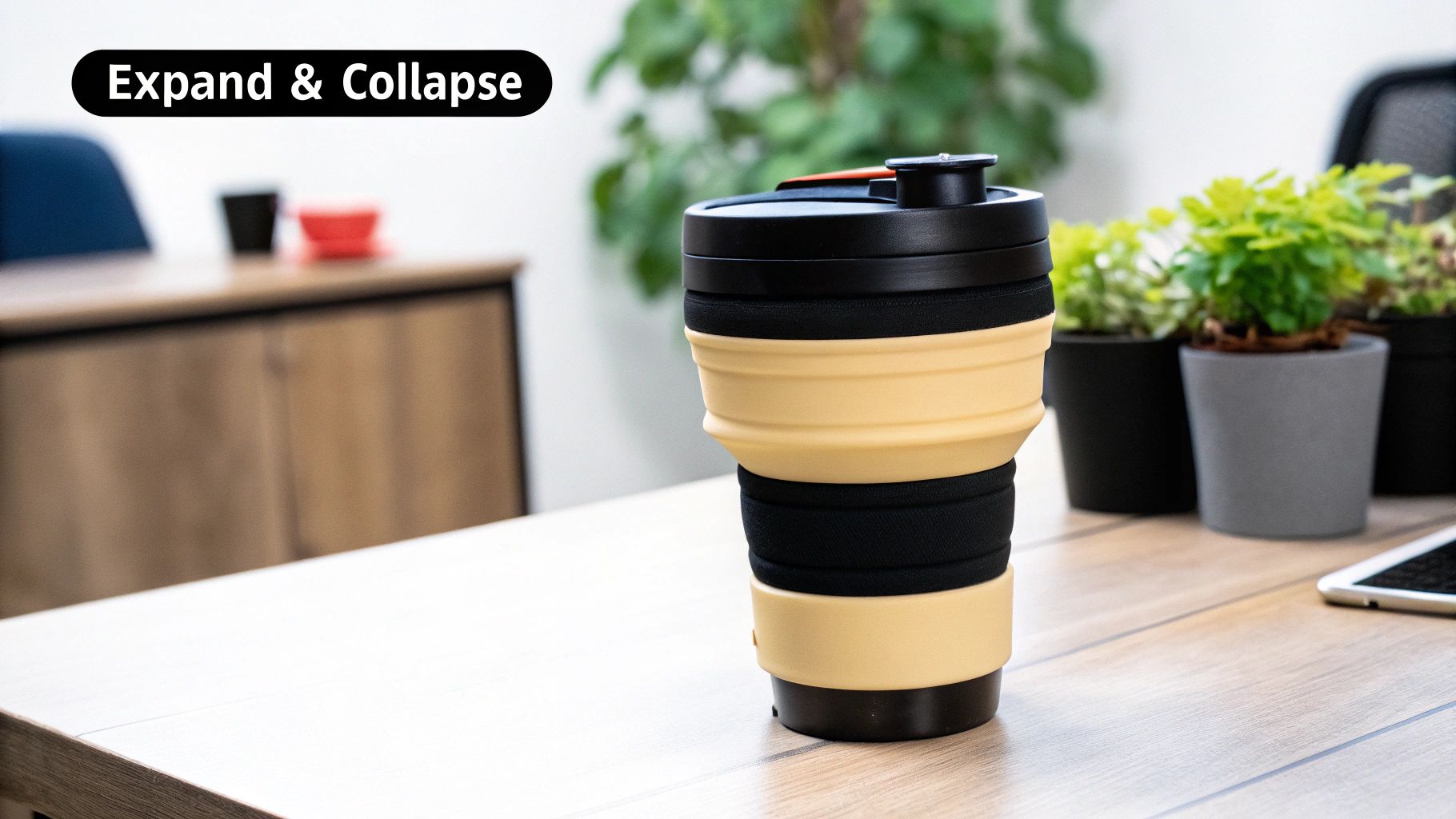 A beige and black collapsible mug stands on a wooden desk, demonstrating its expand and collapse feature.