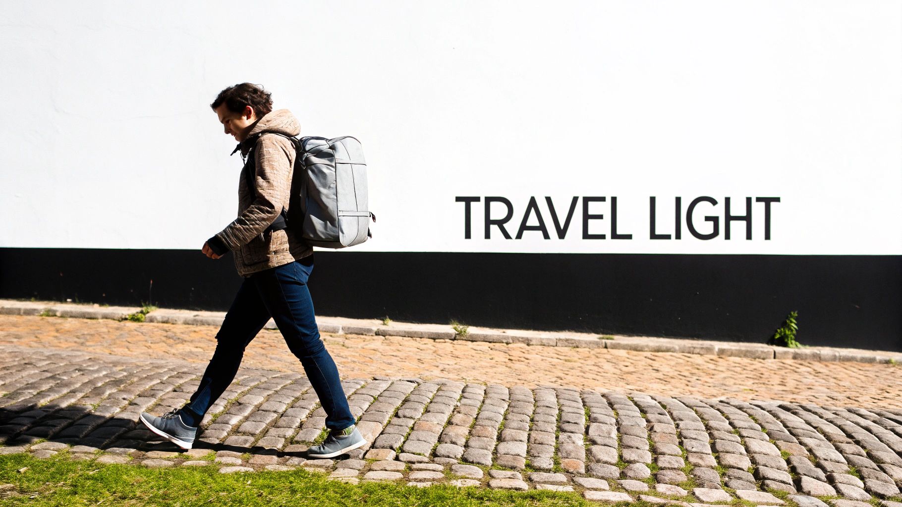 A person with a backpack walks on a cobblestone path past a wall with 'TRAVEL LIGHT' written on it.