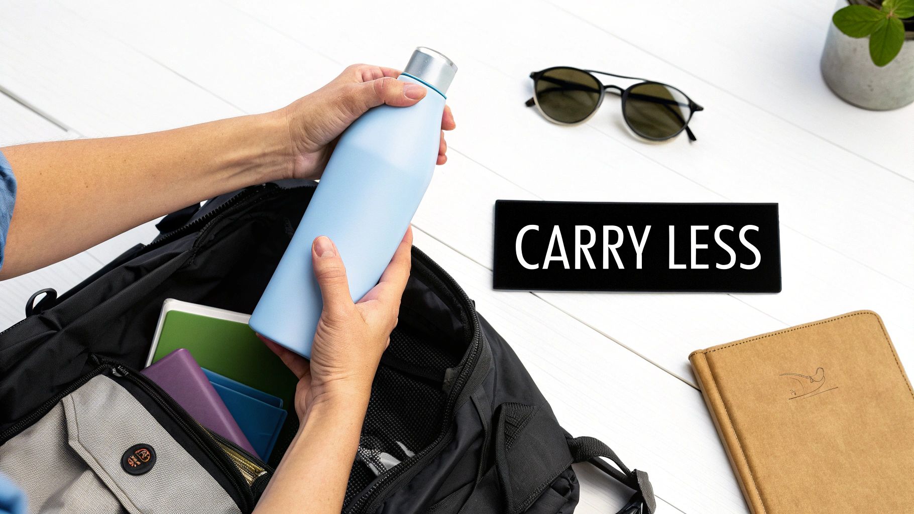Person putting a light blue reusable water bottle into a black backpack with books. A 'CARRY LESS' sign is on the white table.
