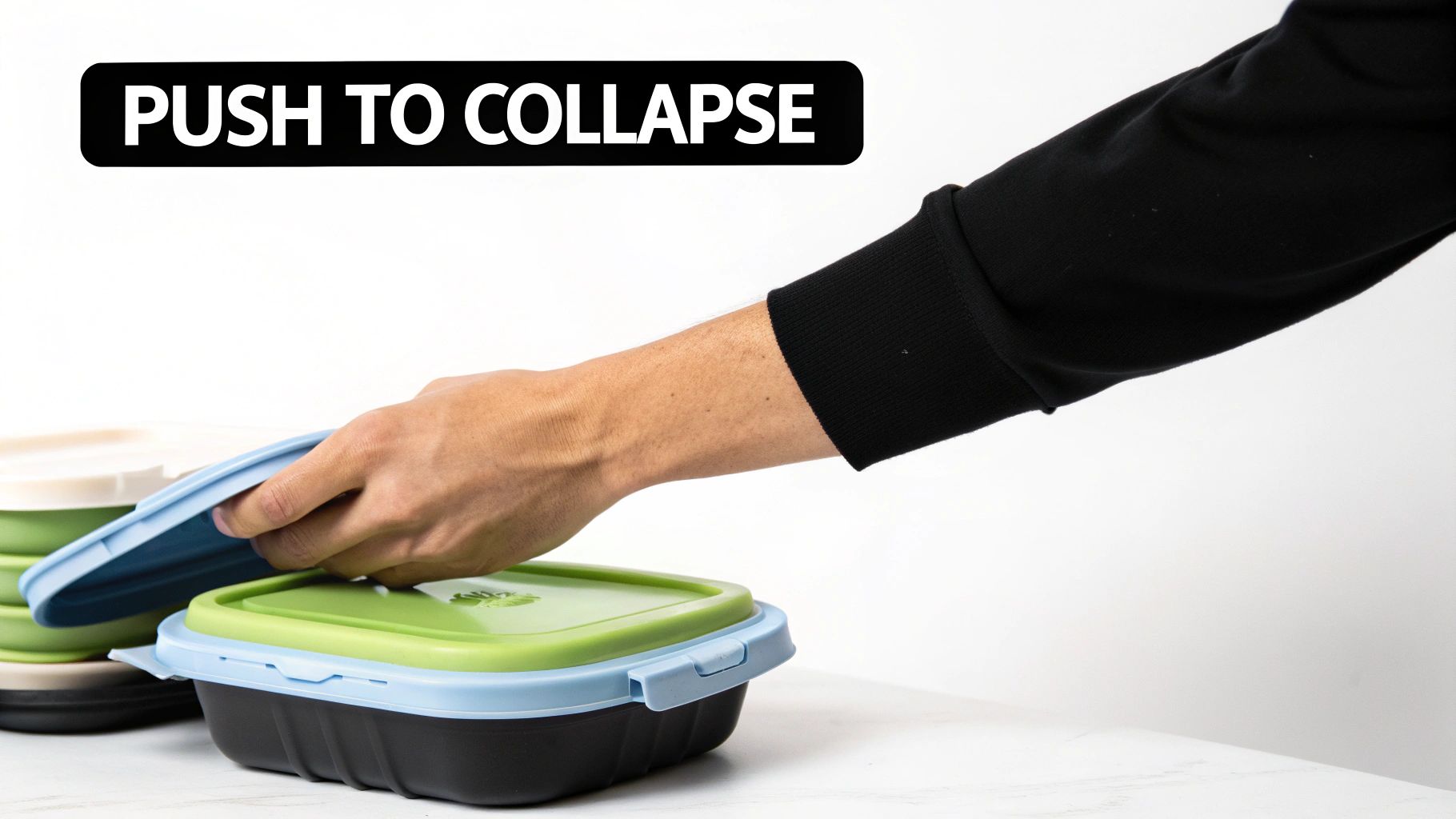 A hand pushes down on a blue lid to collapse a green and black silicone food storage container.