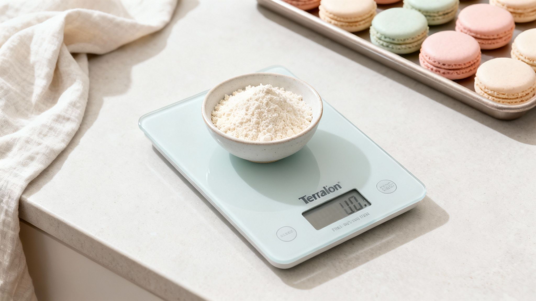 Une balance de cuisine Terralon affiche 0.0 avec un bol de poudre blanche. Un plateau de macarons colorés est en arrière-plan.