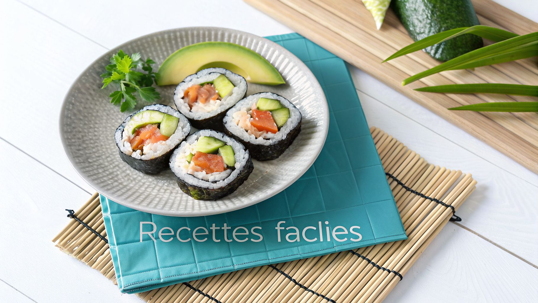 Sushi maki saumon avocat concombre sur une assiette grise, avec une natte de sushi et un tapis turquoise.