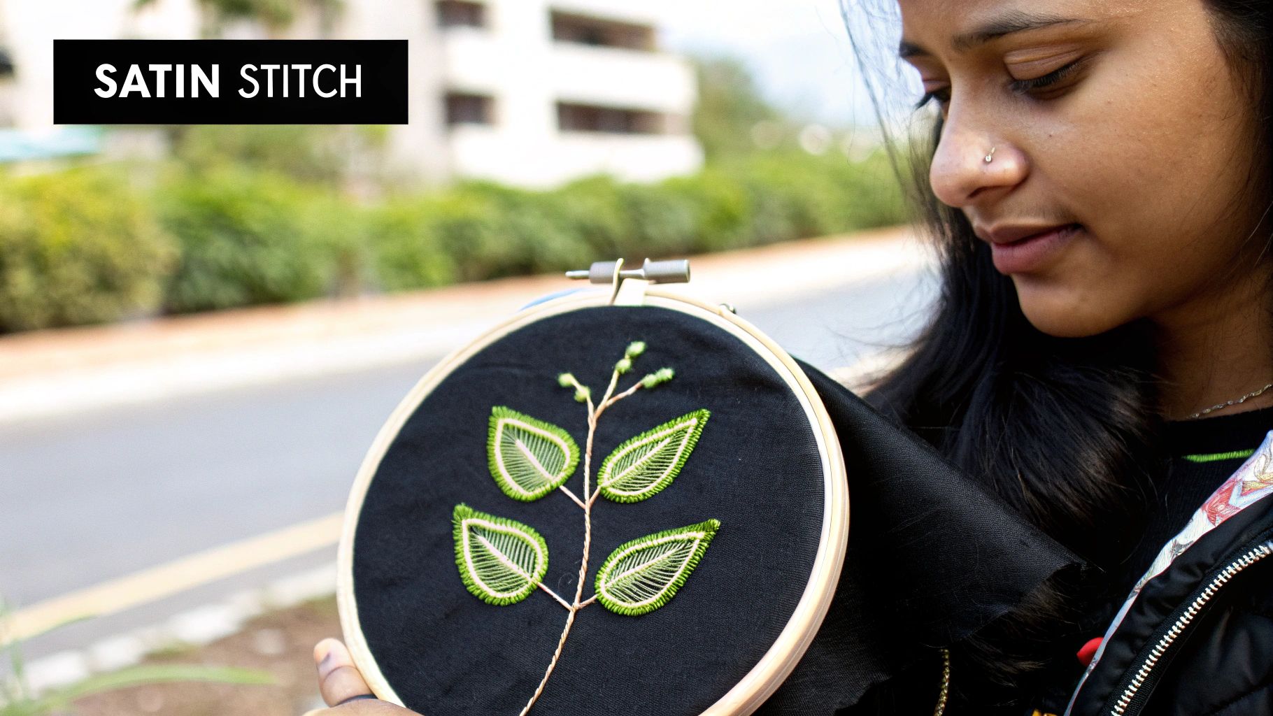 A person holds an embroidery hoop with a black fabric featuring a green satin stitch leaf design.