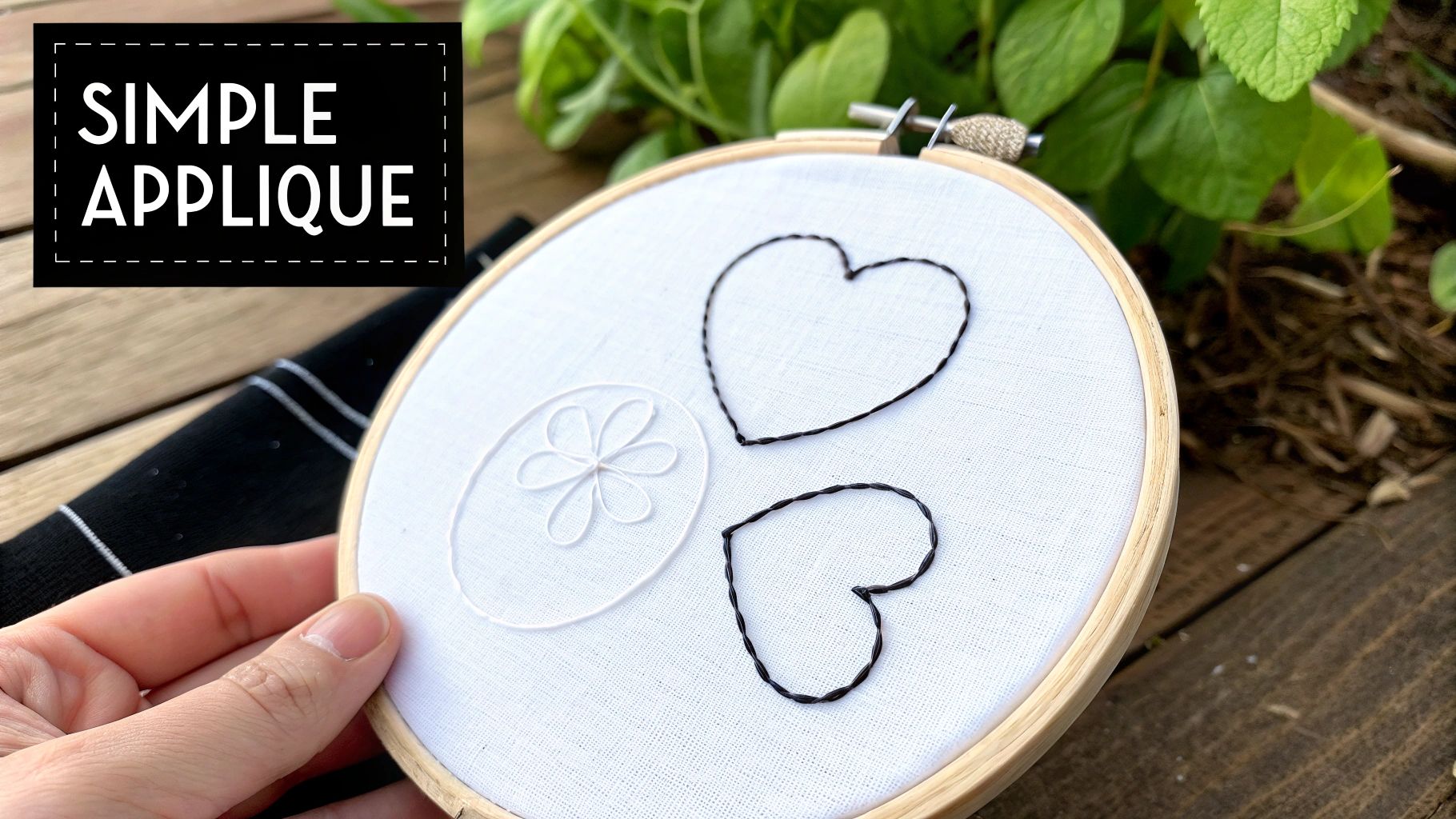 A hand holds an embroidery hoop with simple applique designs of black hearts and a white flower on white fabric.