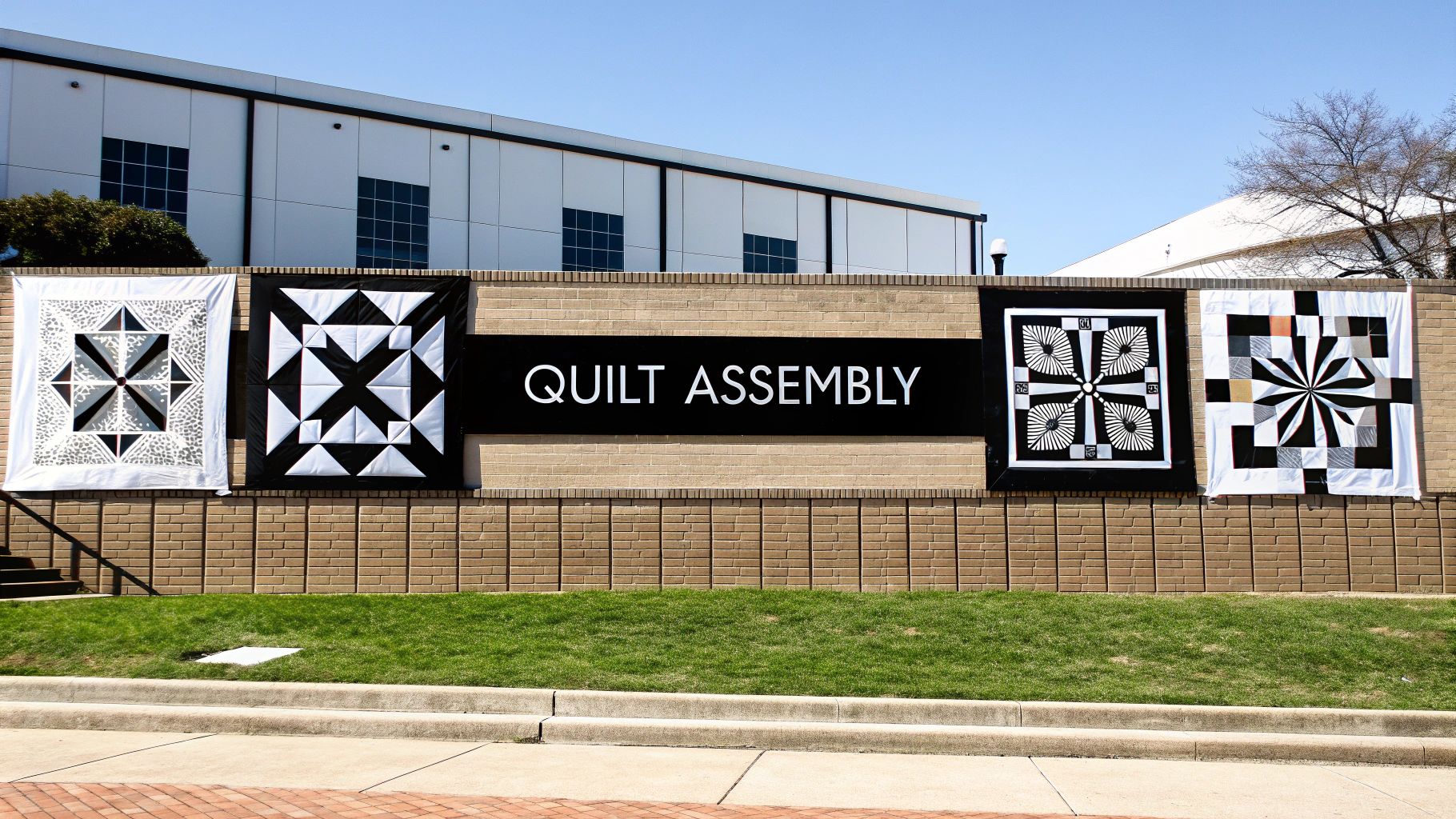 Five unique black and white patterned quilts and a 'QUILT ASSEMBLY' banner displayed on an outdoor brick wall.