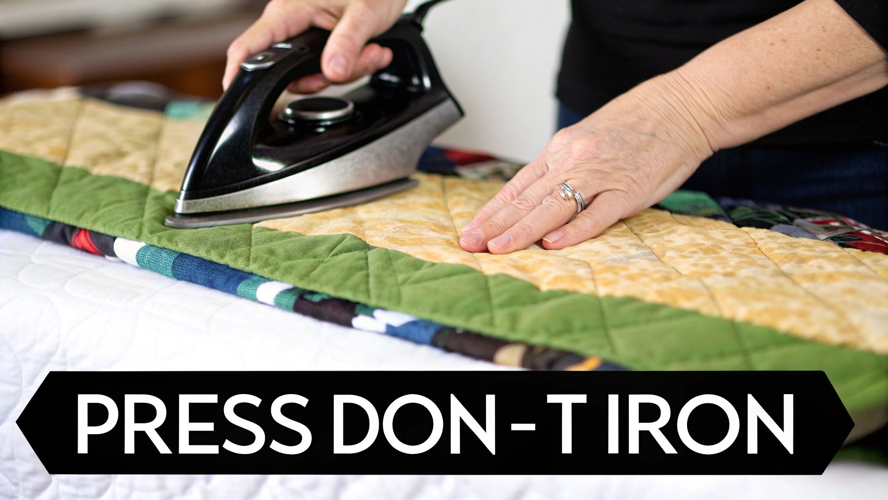 Hands pressing a colorful quilted fabric with a black iron on a white ironing board.