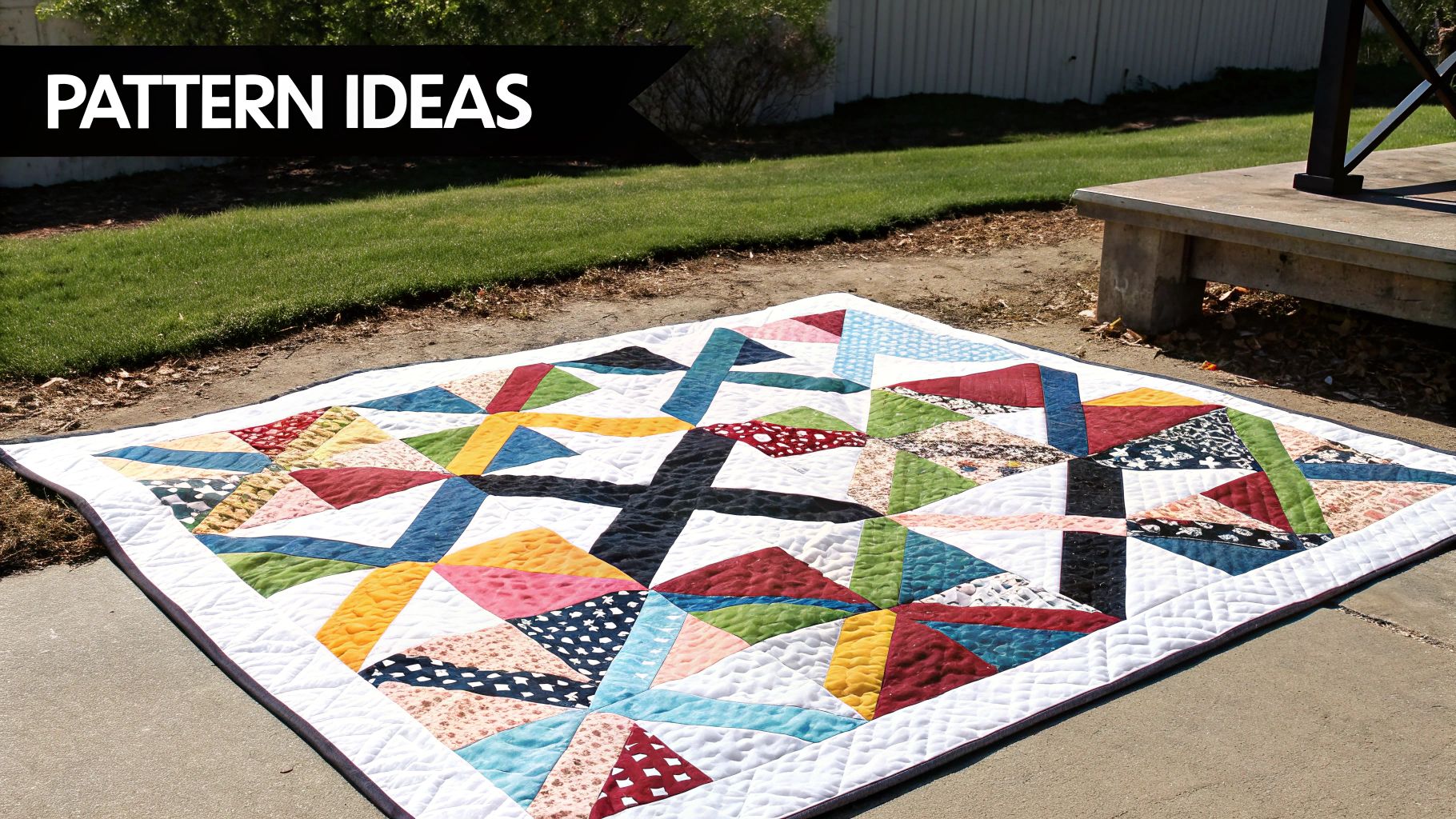 A vibrant, colorful geometric quilt with various patterns laid on a concrete path next to green grass.