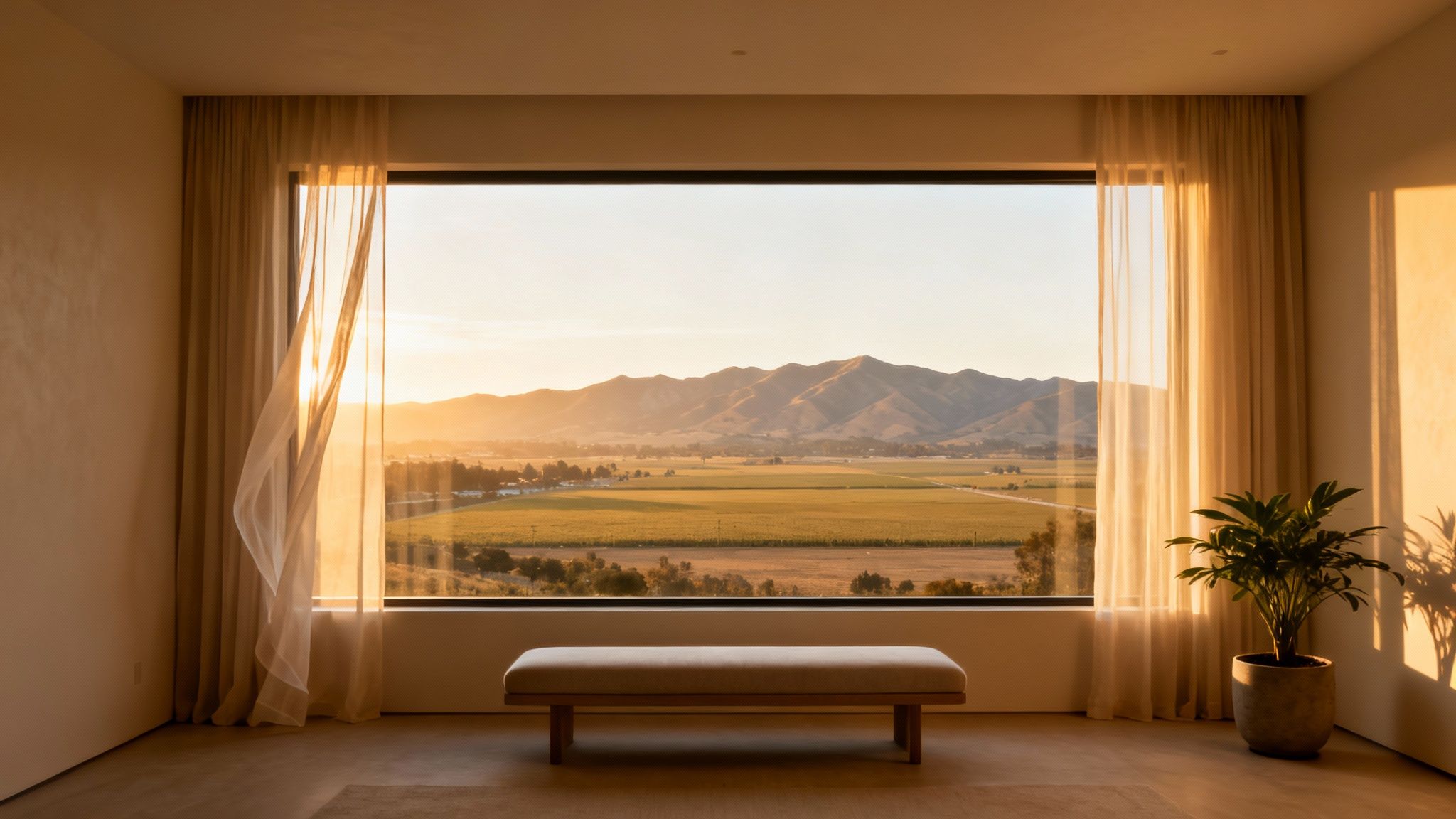 A large window overlooks a golden valley with mountains and fields during a beautiful sunset.