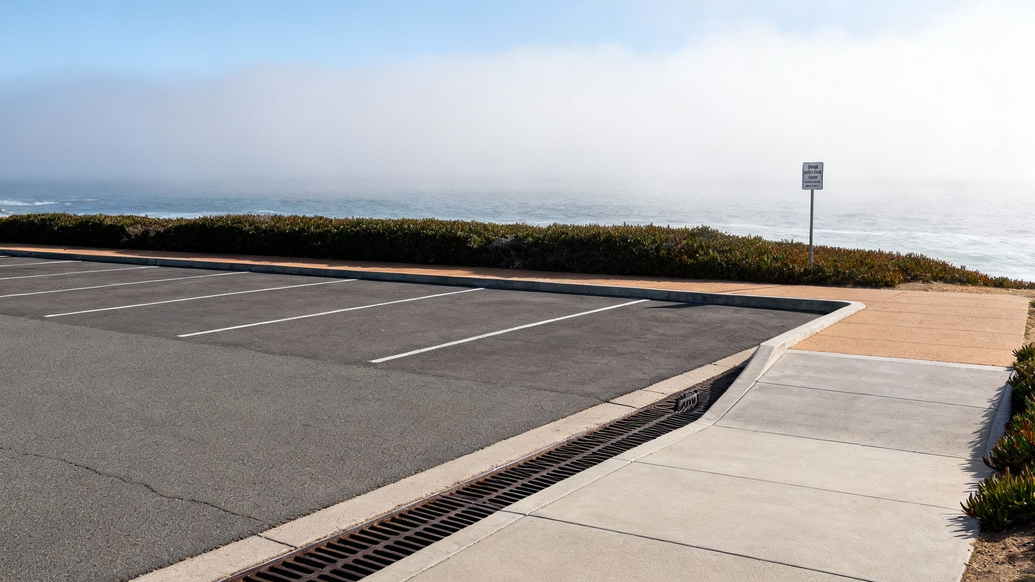 Empty asphalt parking lot with white lines overlooking a foggy ocean and coastal vegetation.