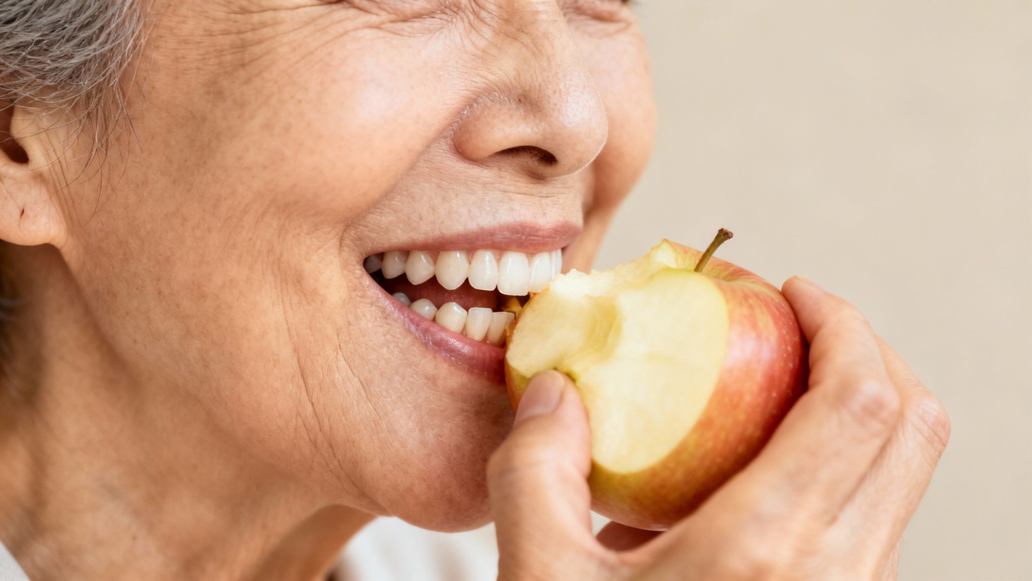 Close-up of an older woman happily biting into a fresh red apple, showing her healthy teeth.