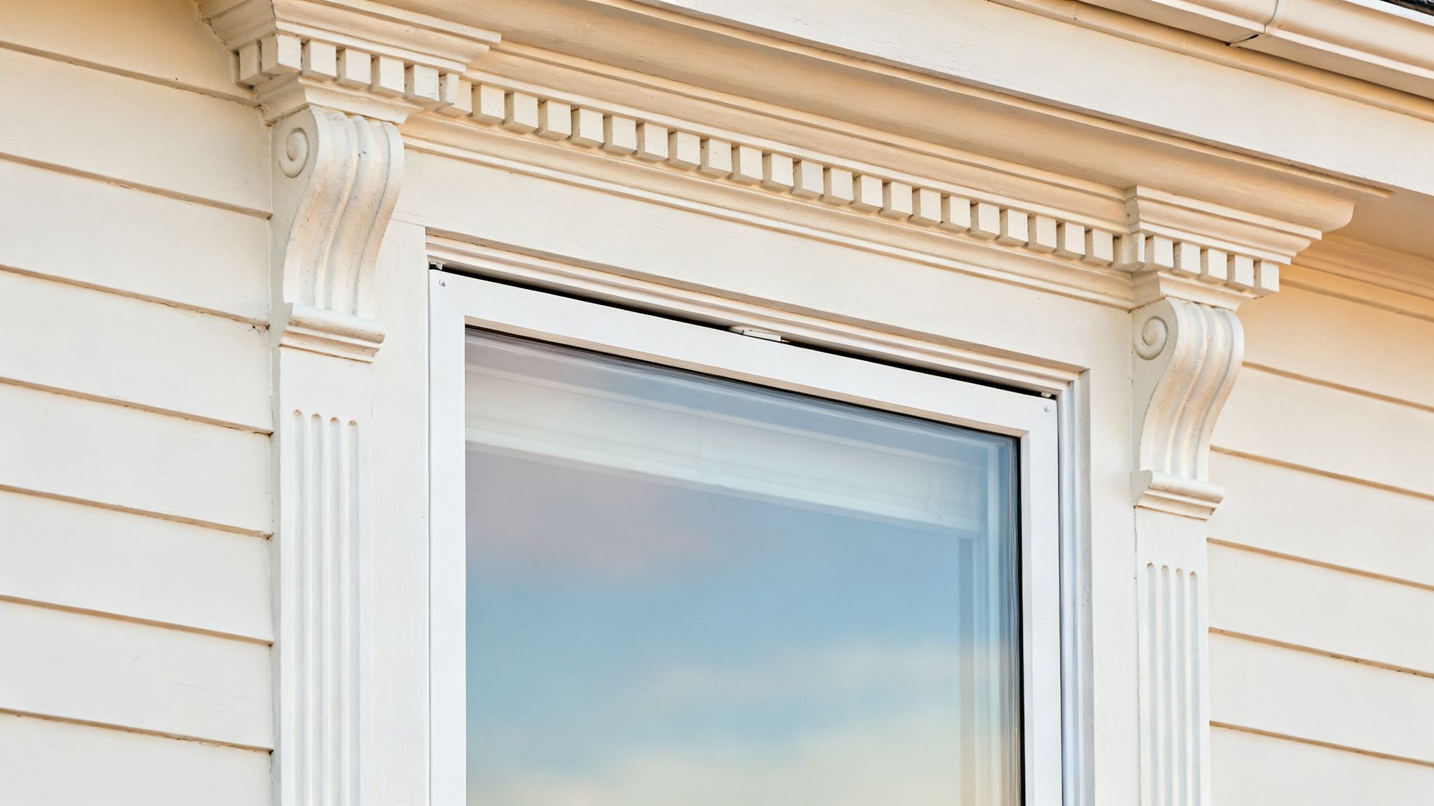 Detail of a white window frame with classical decorative trim, corbels, and siding.