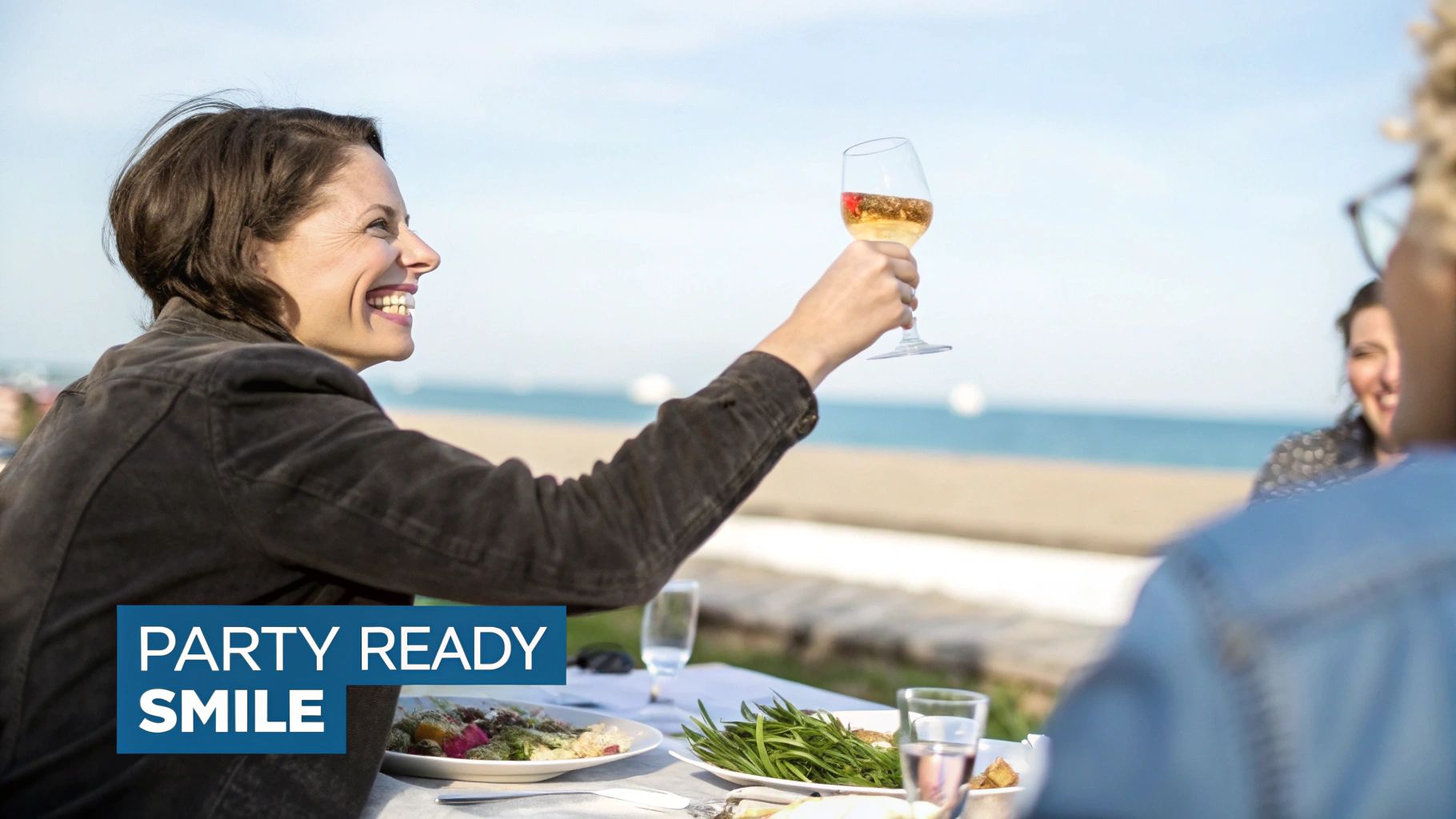 A smiling woman raises a glass of white wine, toasting with friends at an outdoor beachside gathering.