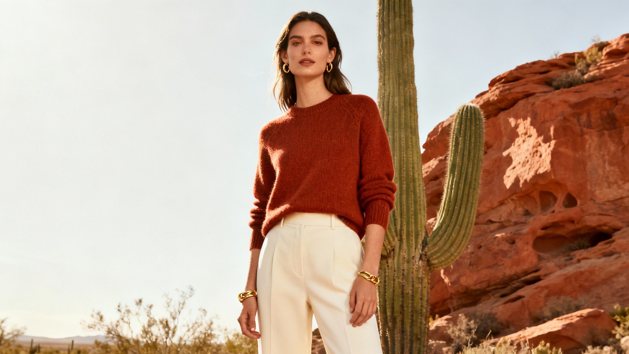 Woman in rust sweater and white pants posing outdoors near desert cactus and red rocks