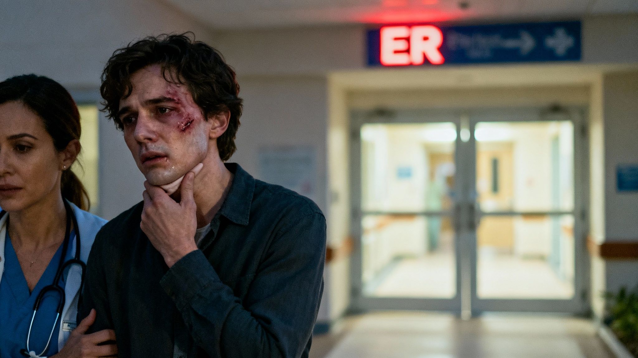 A distressed man with facial injuries touches his throat next to a concerned doctor in a hospital ER.