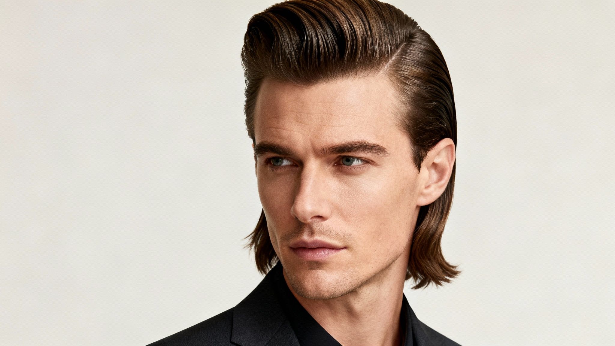 Hombre con peinado clásico y brillante, cabello castaño peinado hacia atrás, mirando de lado en un traje oscuro.
