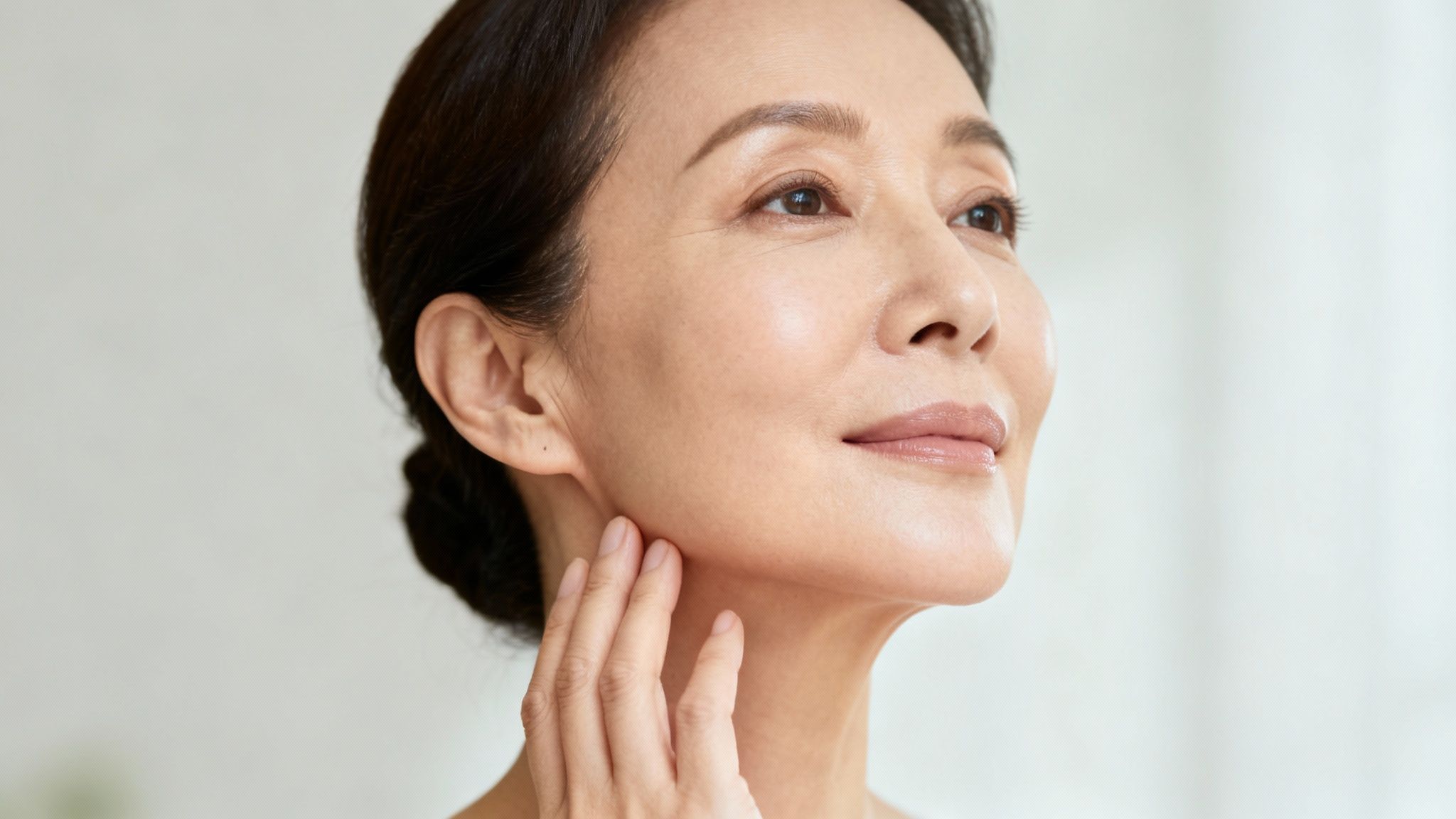 Primer plano de una mujer asiática madura sonriendo, mostrando piel radiante mientras toca su mandíbula.