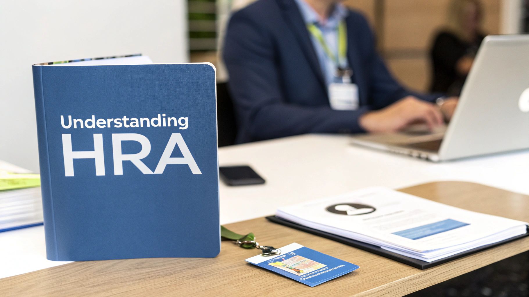 A blue book titled 'Understanding HRA' on a wooden desk, with a person working on a laptop in the background.