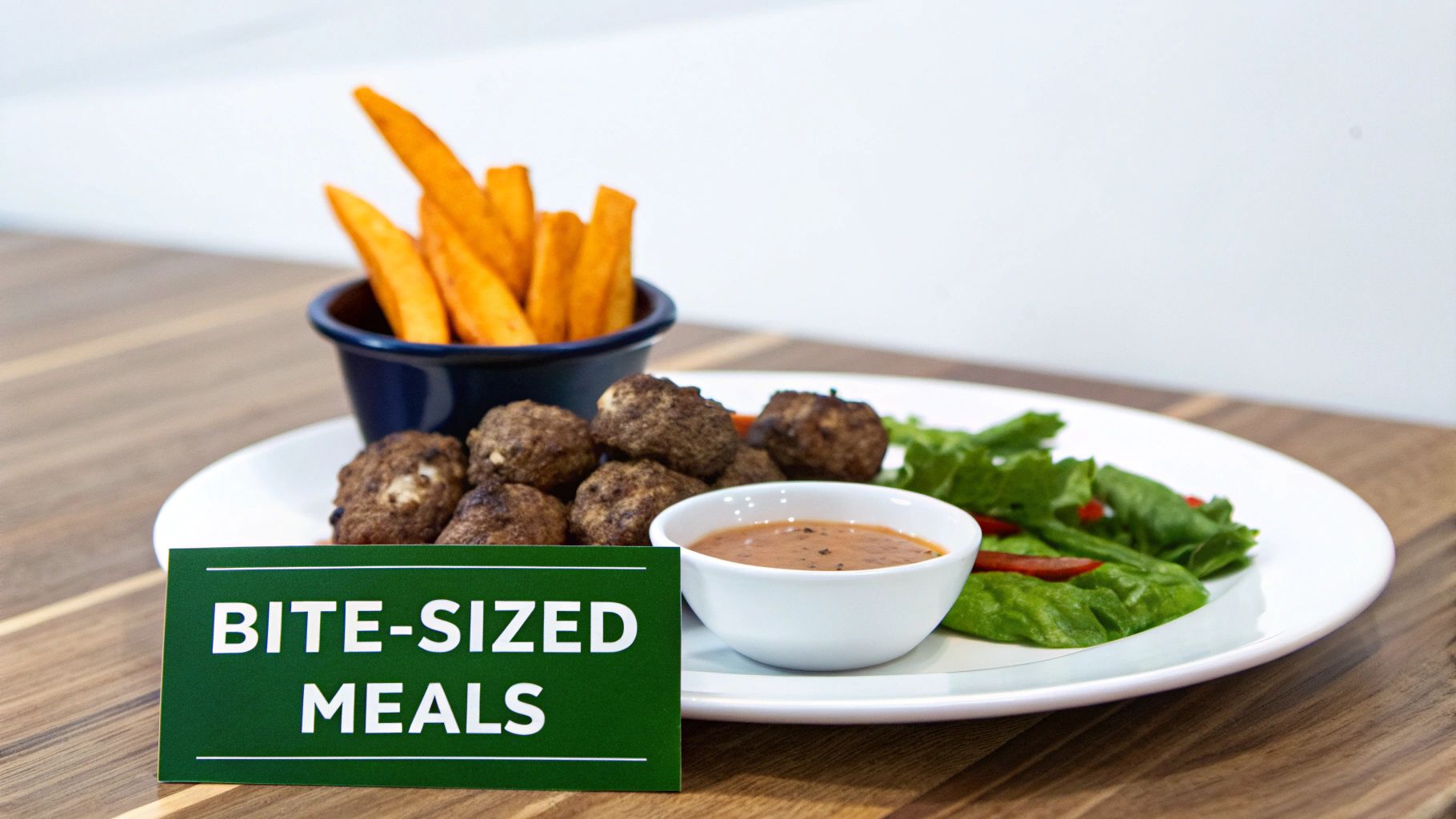 A plate of bite-sized meatballs, sweet potato fries, salad, and sauce on a wooden table.