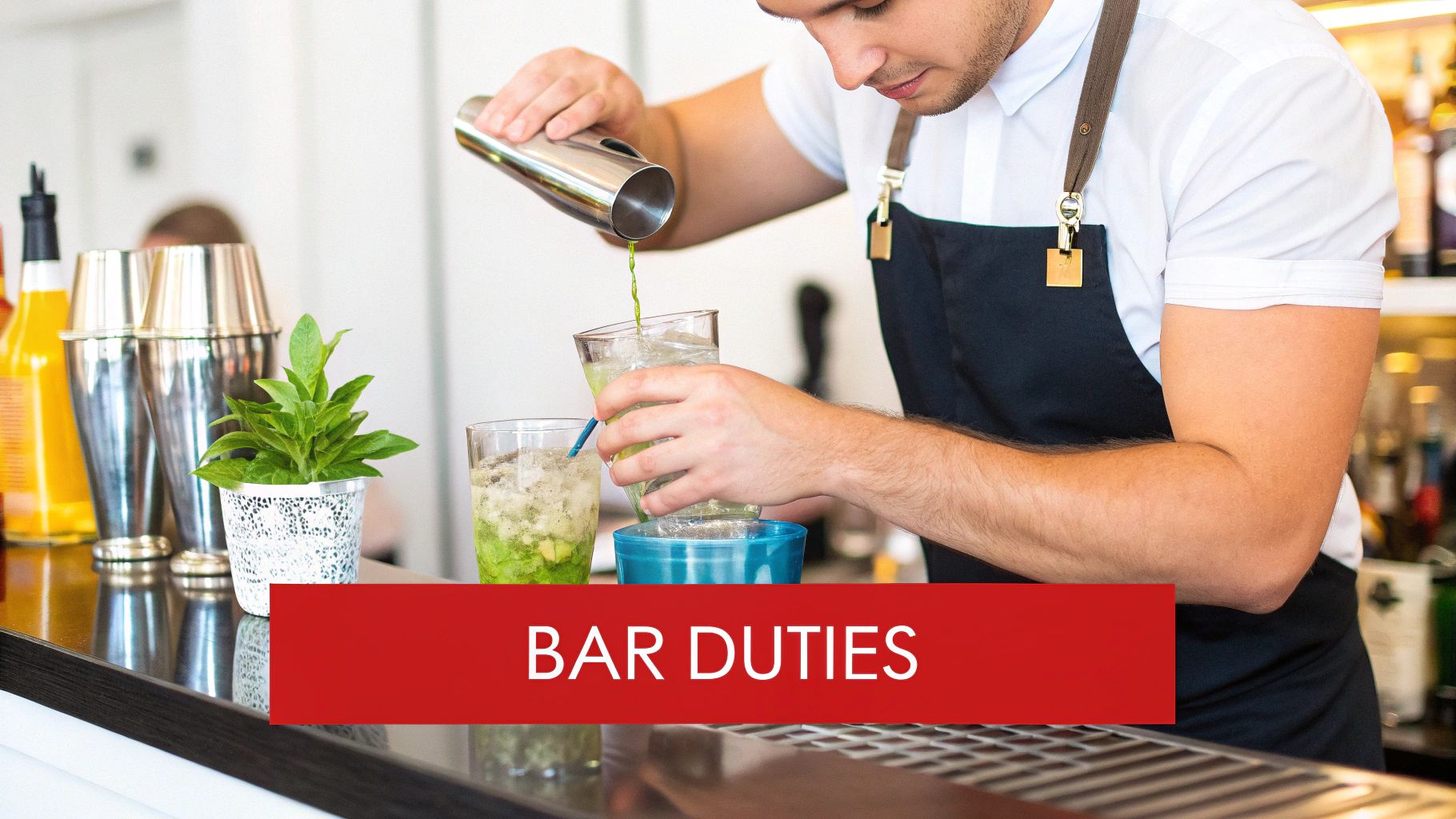 A male bartender in an apron expertly pours a green drink from a shaker into a glass on a bar.
