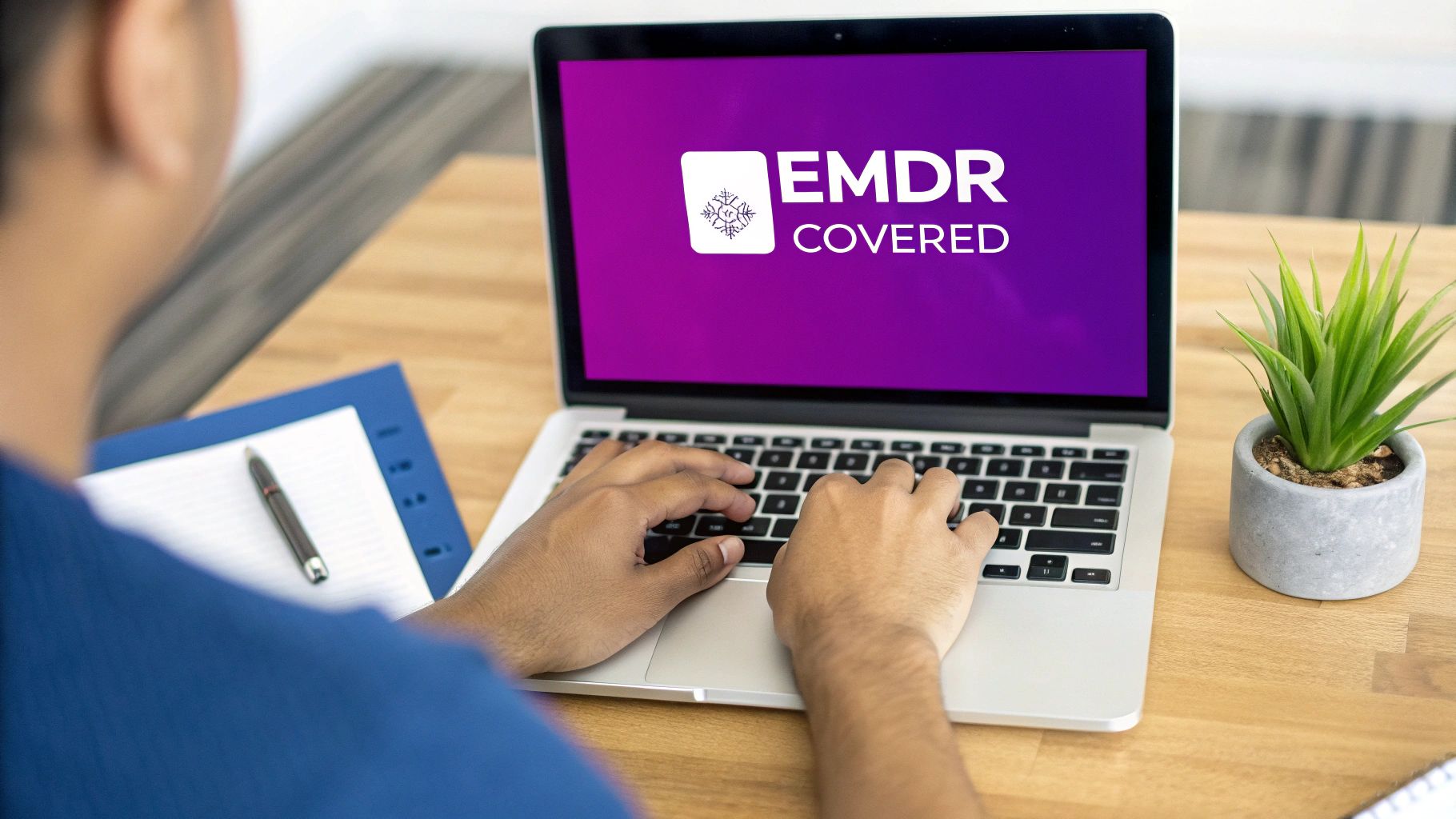A person types on a laptop displaying 'EMDR COVERED' on a vibrant purple screen, on a wooden desk.