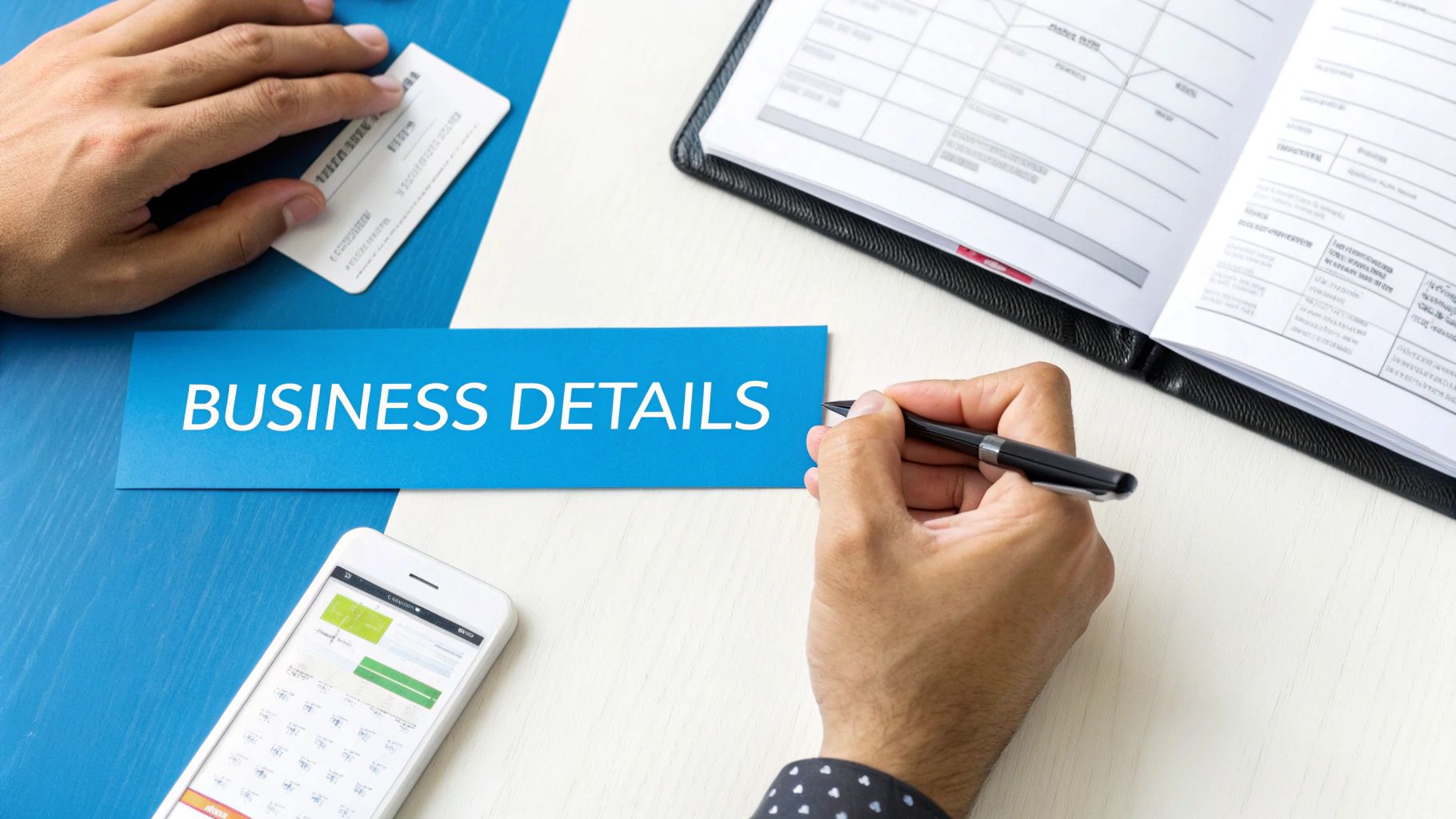 Hands on a desk with a 'BUSINESS DETAILS' sign, card, pen, phone, and planner.