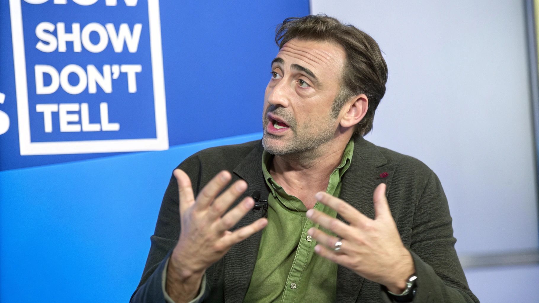 A man in a green shirt and blazer speaks animatedly, gesturing his hands during a presentation.