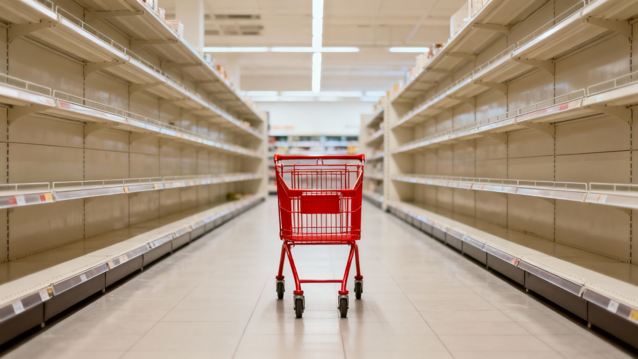 A red shopping cart stands alone in a wide, empty supermarket aisle with completely bare shelves.