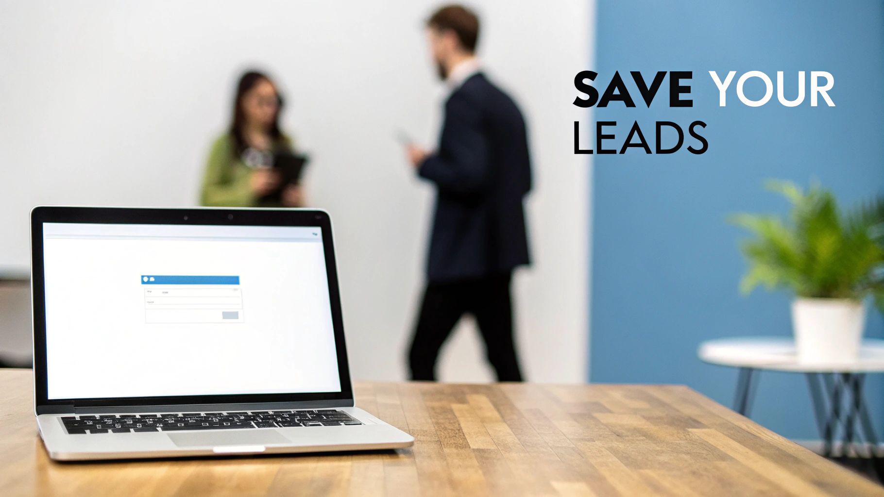 A laptop on a wooden desk displays a form while two people talk in a modern office.