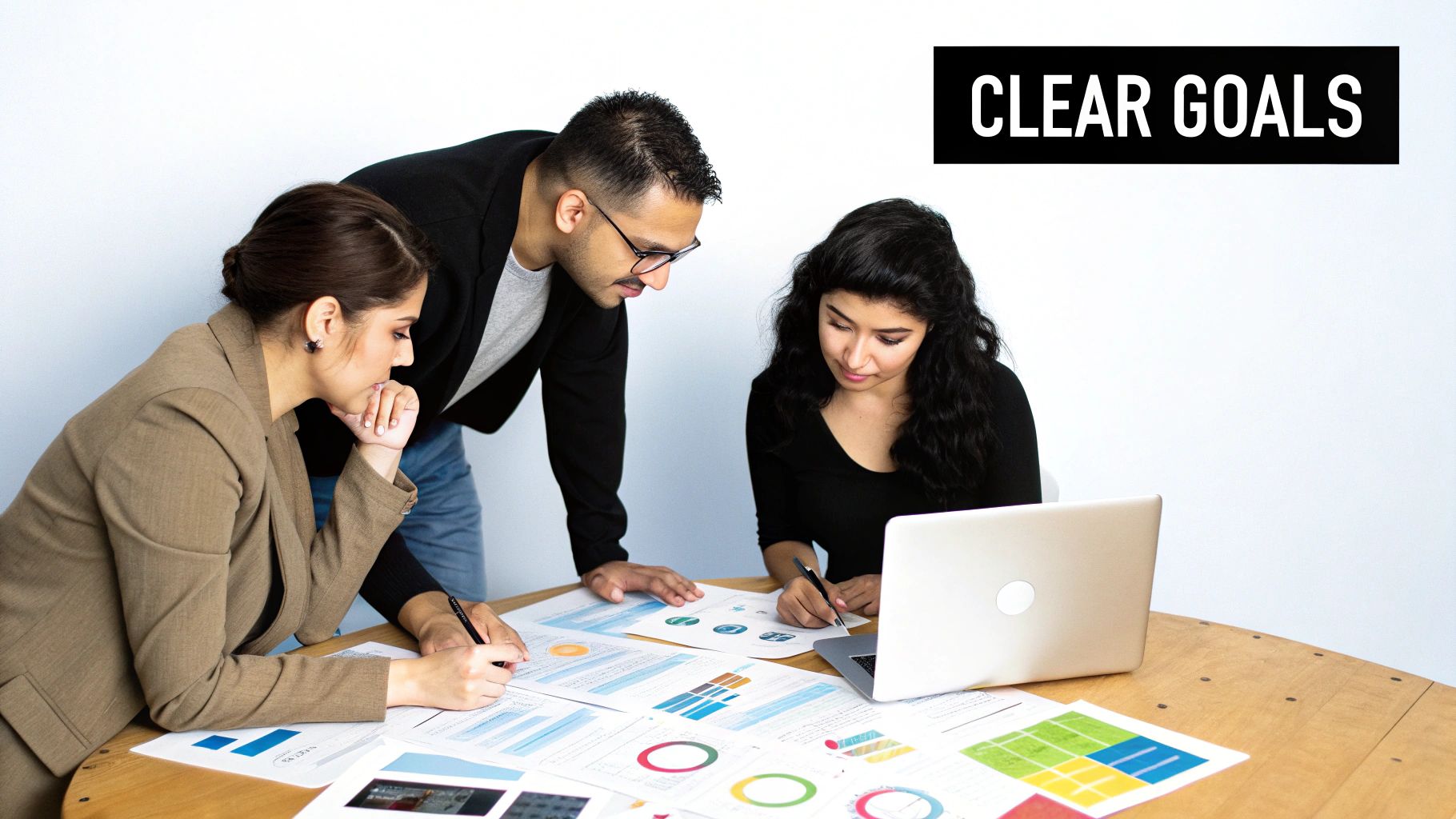 Three colleagues collaborate around a table, reviewing documents and using a laptop, discussing clear goals.