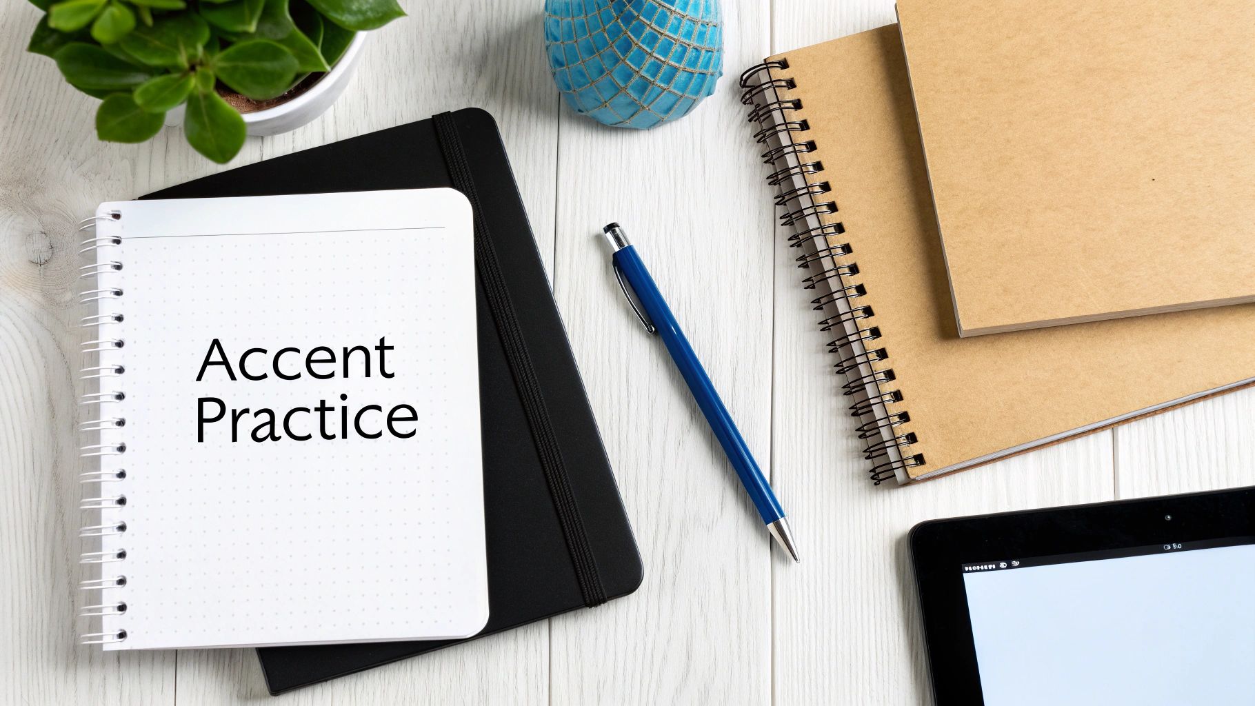 A white spiral notebook with "Accent Practice" written on it, surrounded by a pen, other notebooks, and a plant on a white wooden desk.
