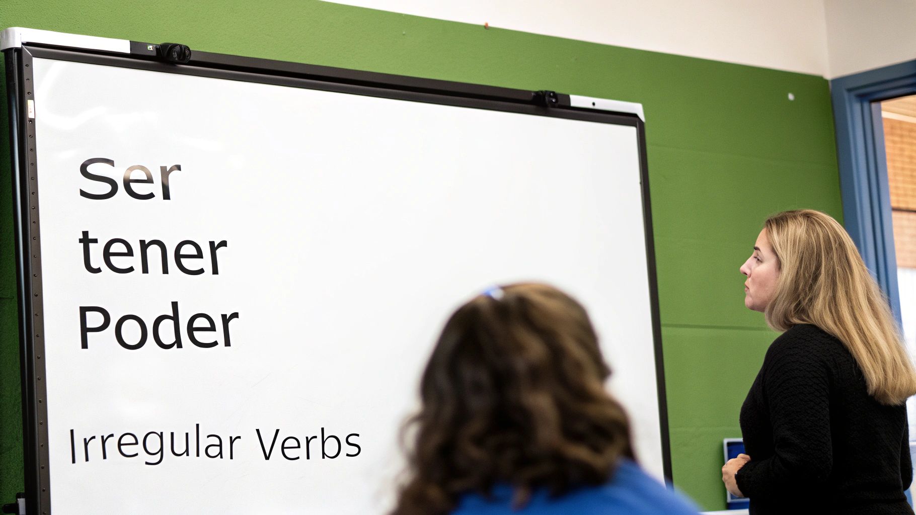 A teacher stands next to a whiteboard displaying Spanish irregular verbs: Ser, tener, and Poder.