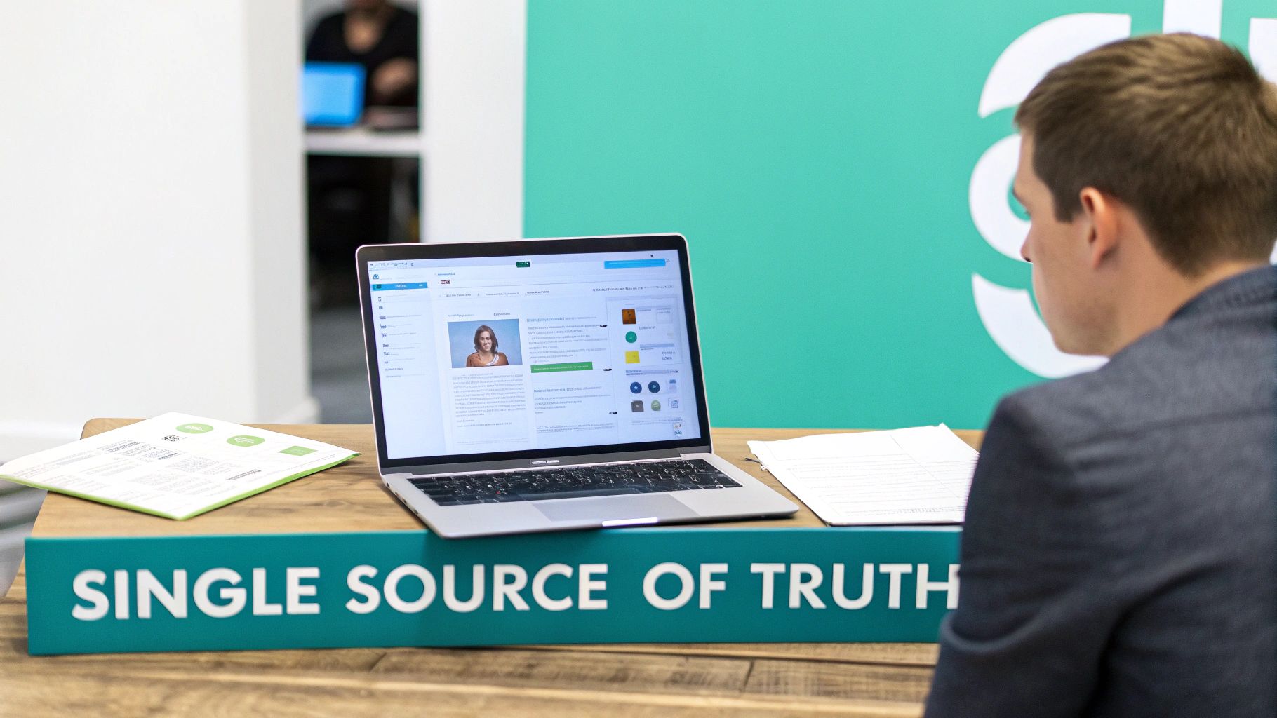 A man focused on a laptop displaying a profile, with a 'SINGLE SOURCE OF TRUTH' sign.