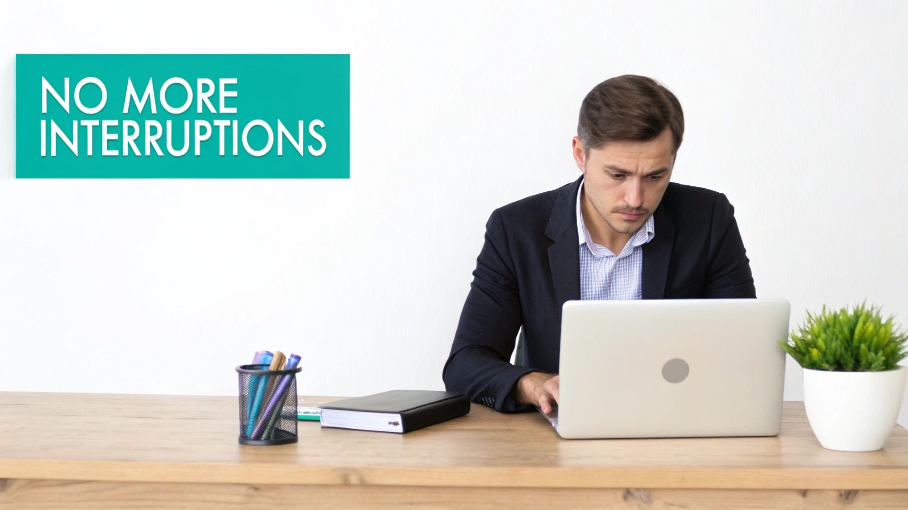 A focused man works on his laptop at a wooden desk, with a sign reading 'NO MORE INTERRUPTIONS'.
