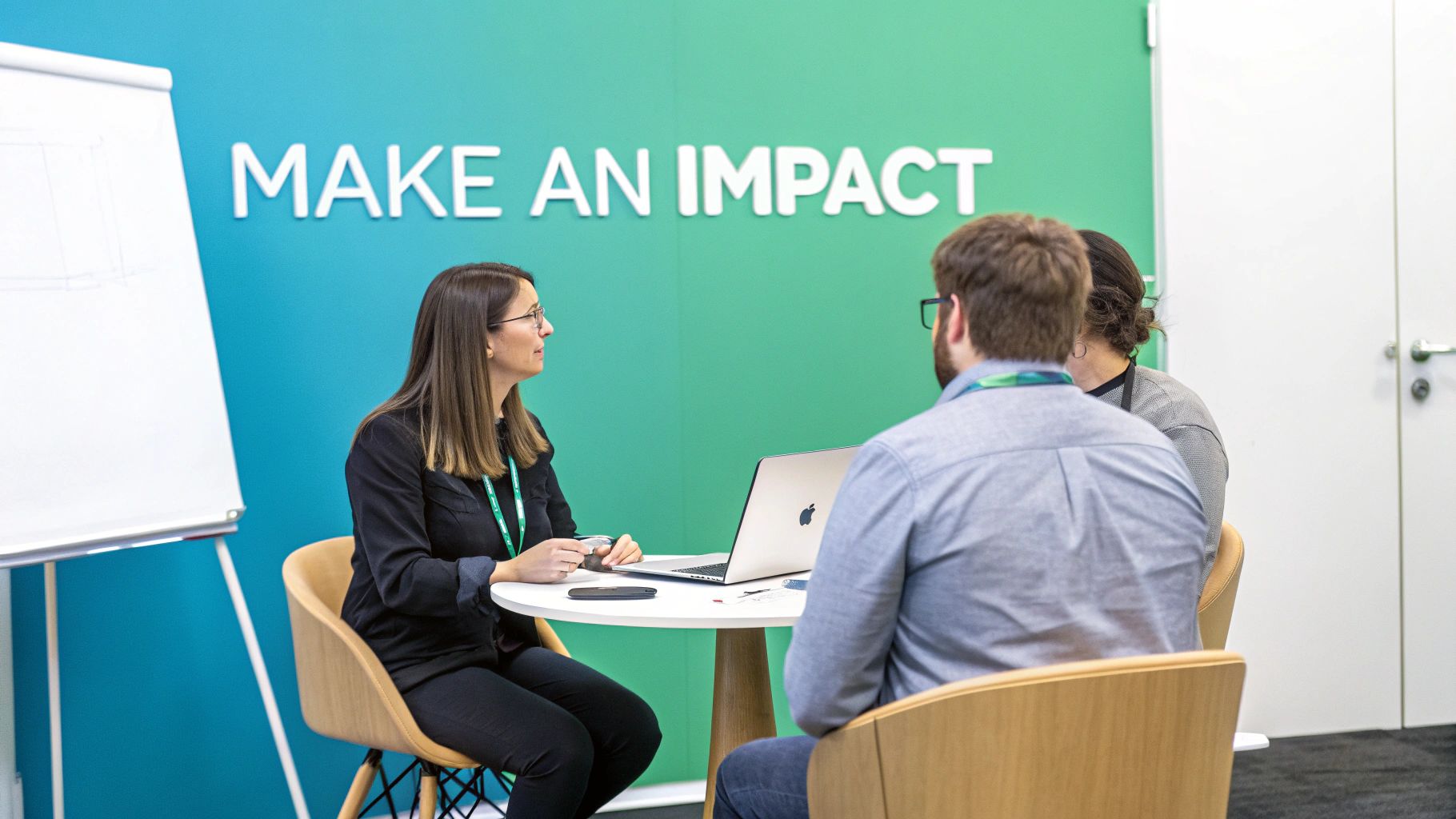 Three professionals discussing in a modern office, one presenting at a table with a laptop.