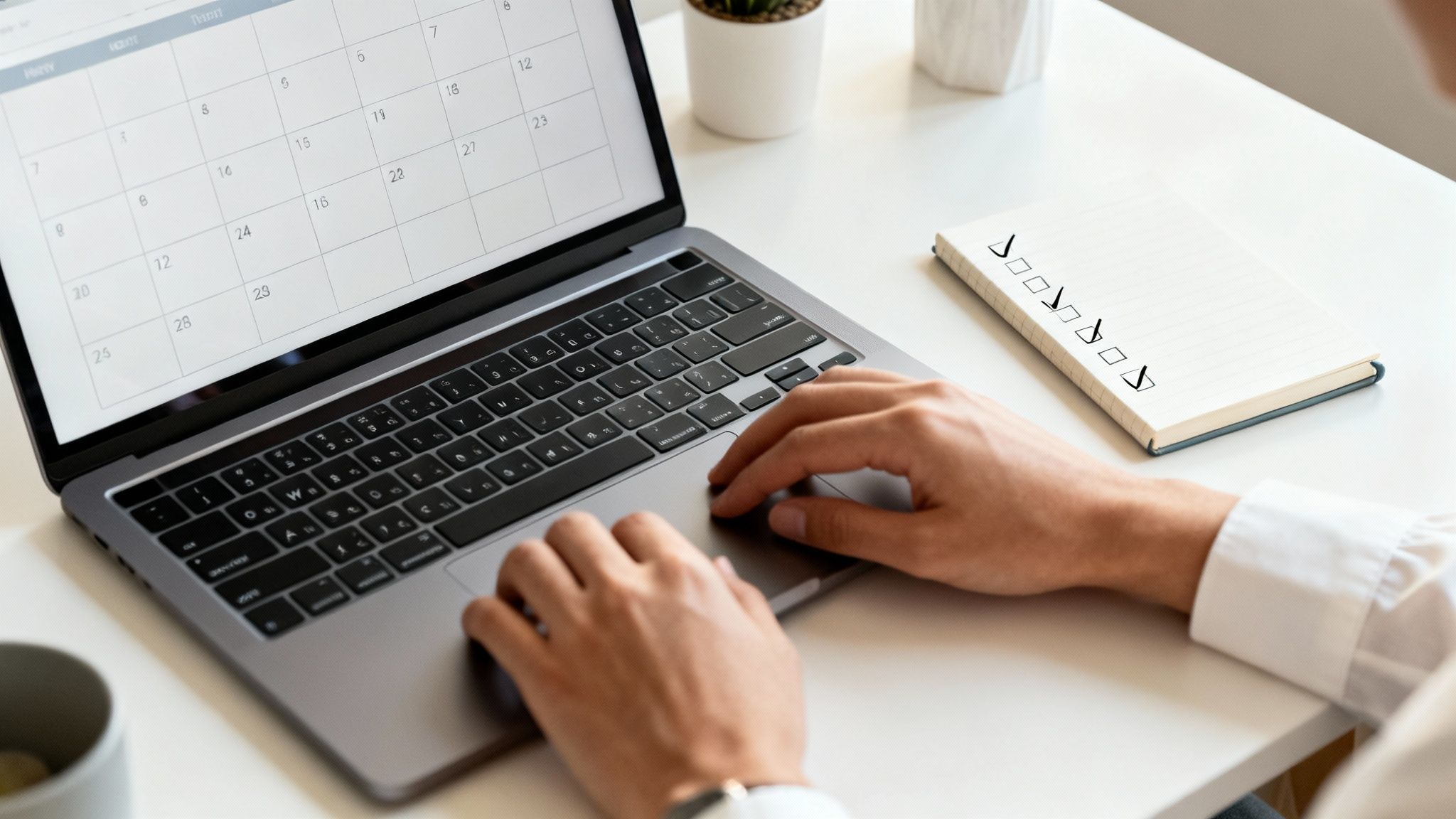 Close-up of hands typing on a laptop with a calendar and a checklist notebook for planning.