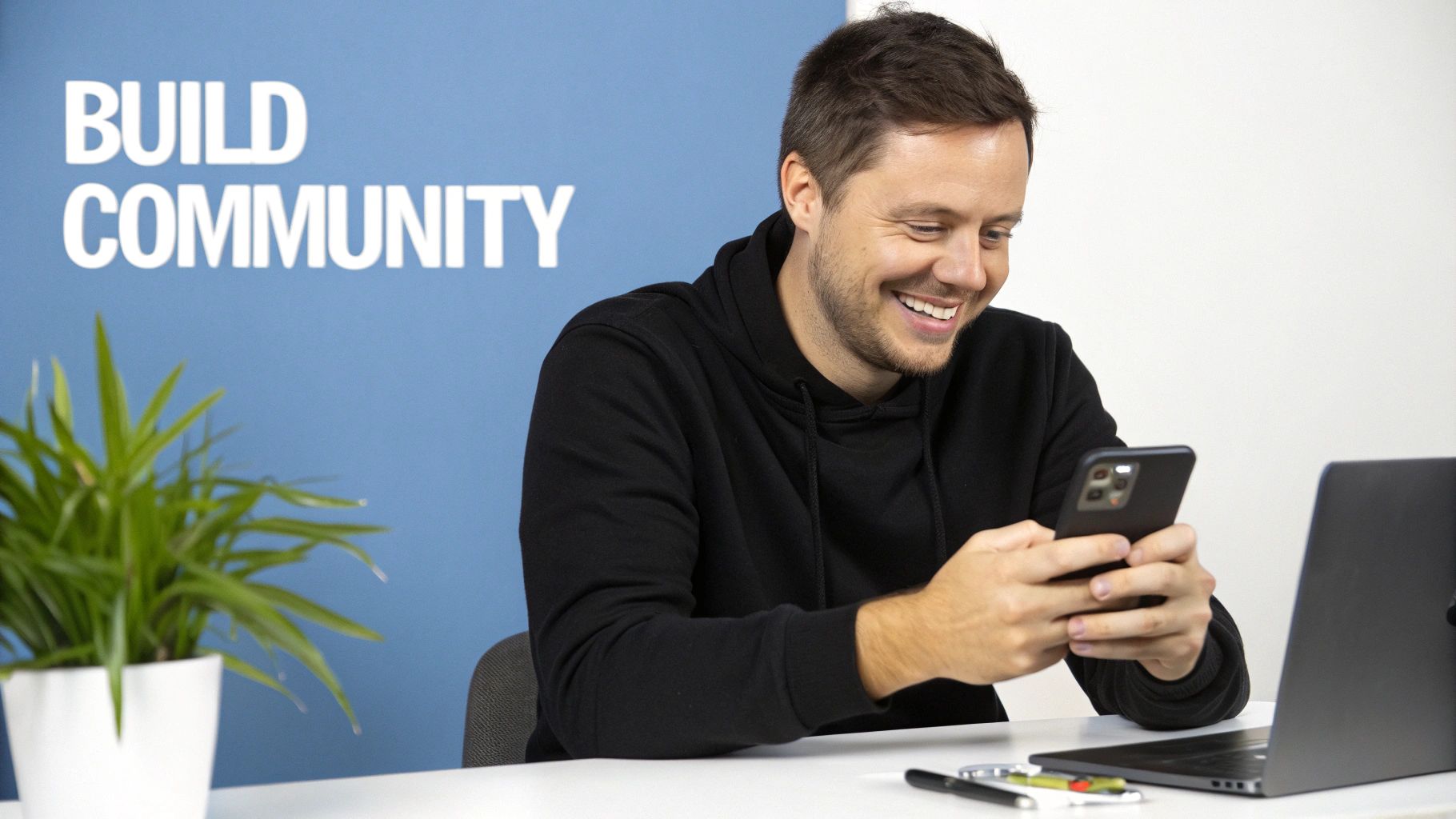 Happy man in black hoodie using a smartphone at a desk with a laptop, plant, and "BUILD COMMUNITY" sign.