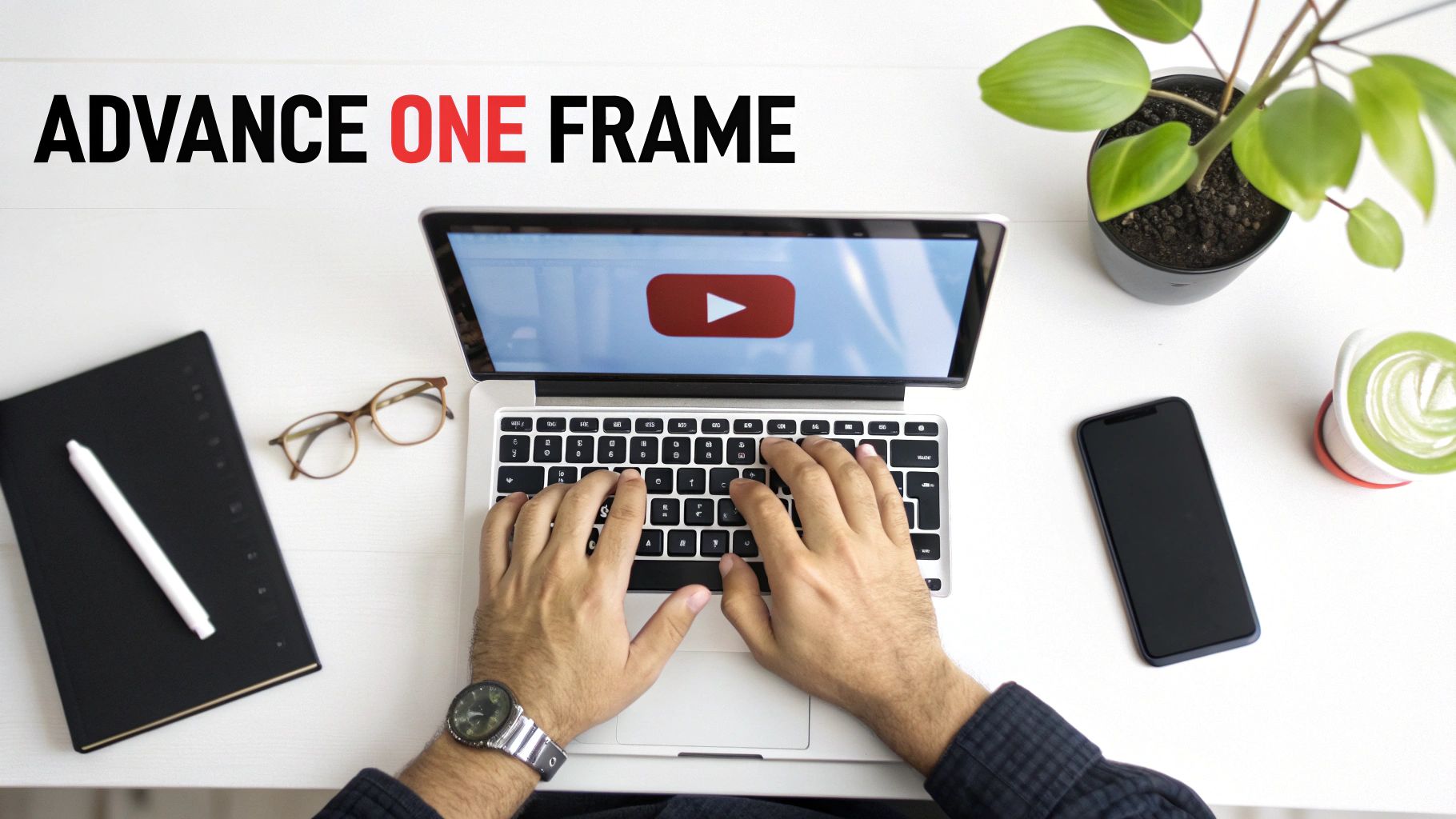 Person typing on laptop displaying YouTube video with workspace including notebook, glasses, phone and plant