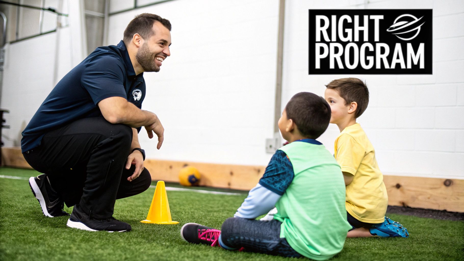 A smiling coach squats on artificial turf, engaging with two young boys sitting in front of him.