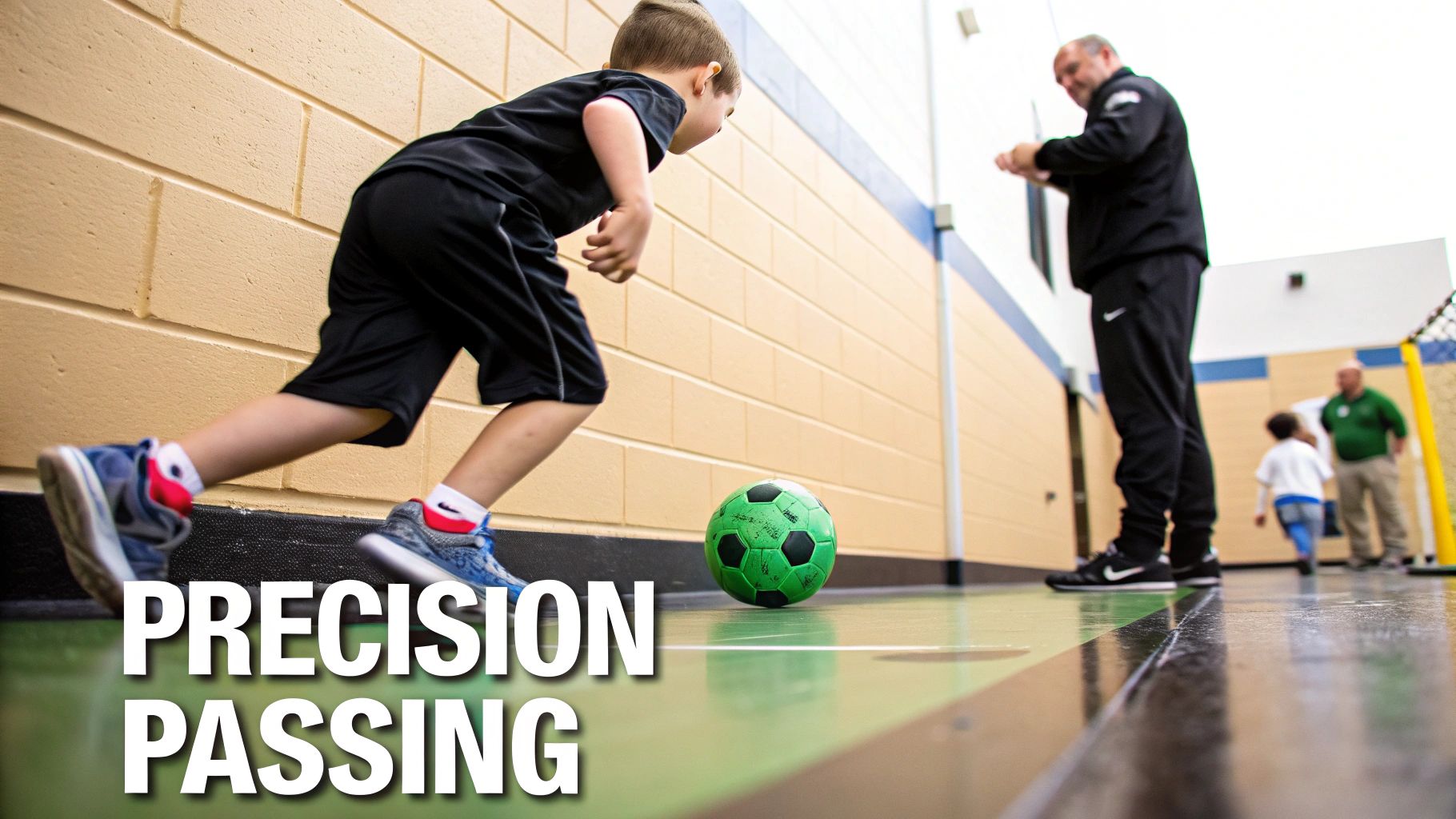 Young boy practices soccer passing with a green ball on an indoor court, observed by a coach.