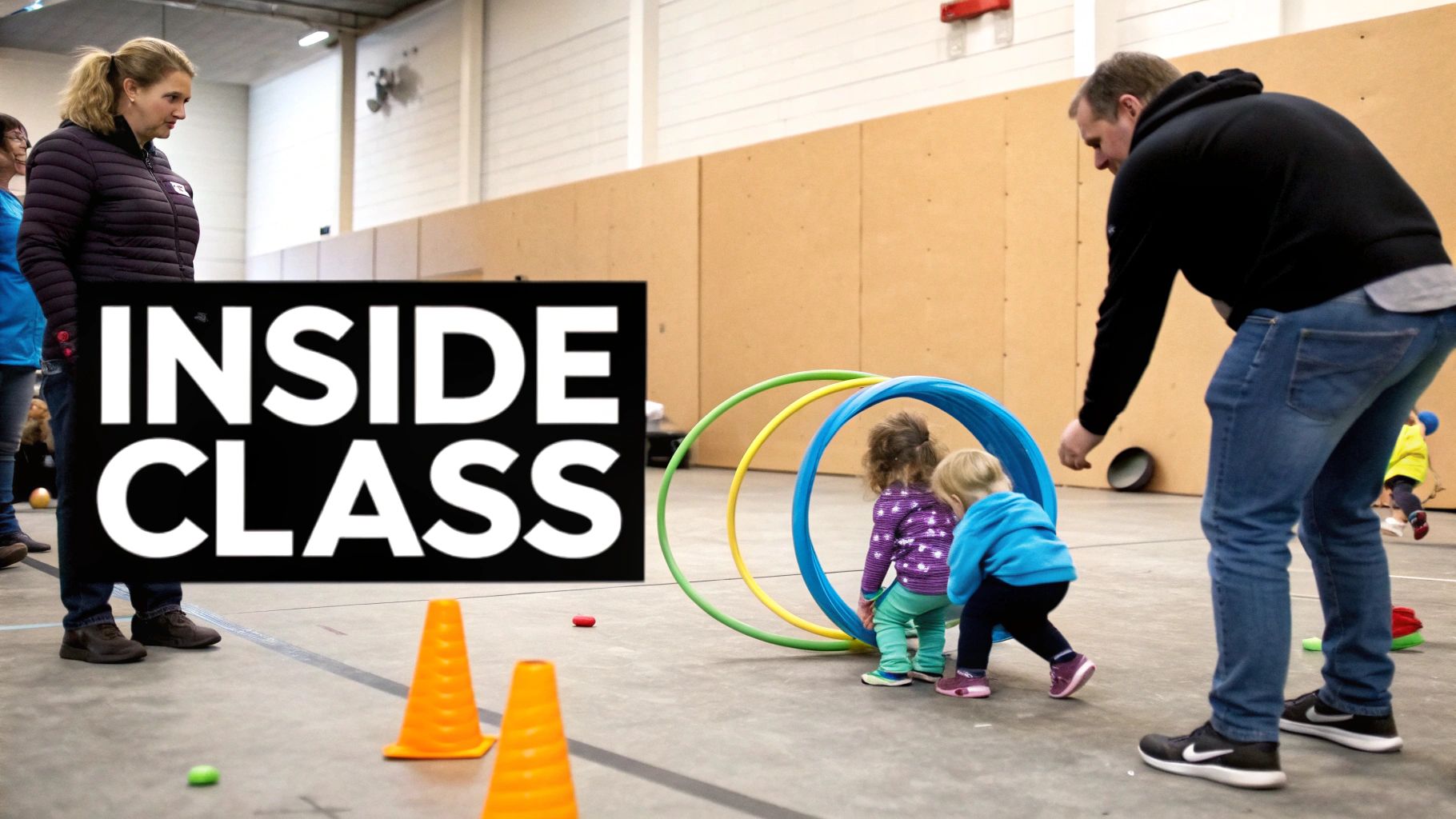 Adults supervise toddlers playing with colorful hoops and cones in an indoor sports class.
