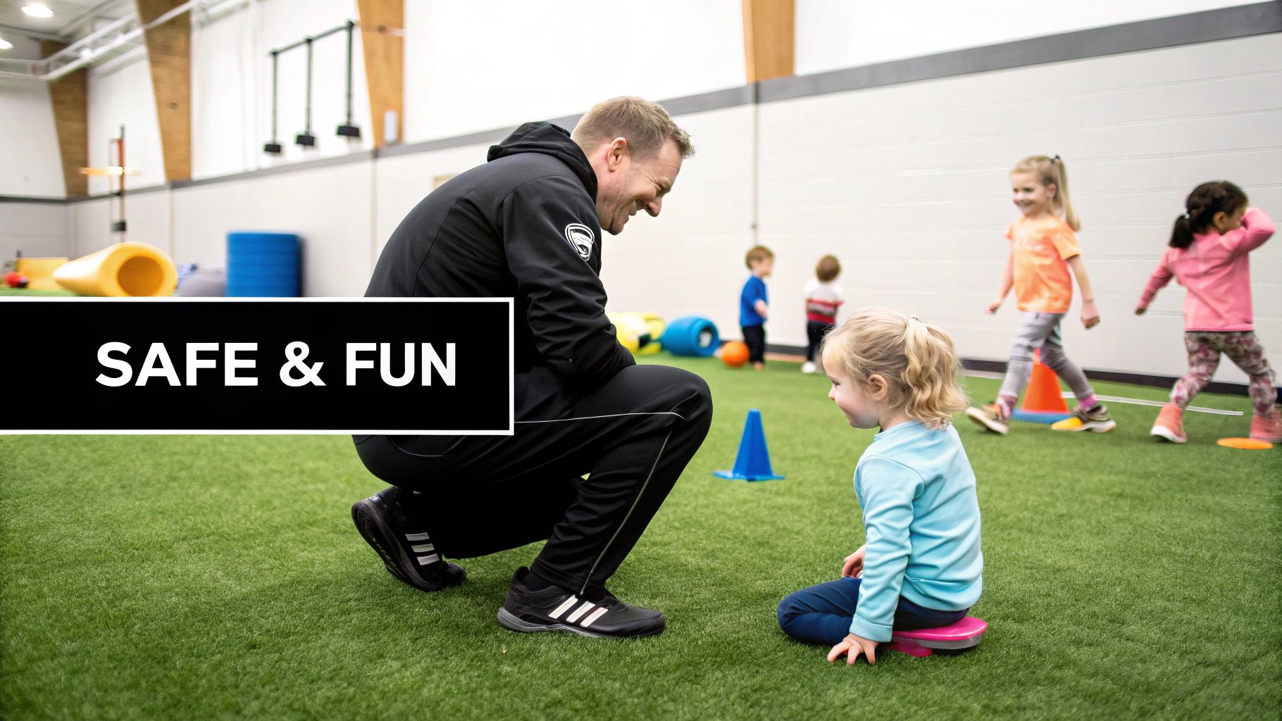 A male coach smiles at a toddler sitting on a pink disc during a fun sports activity on green turf.