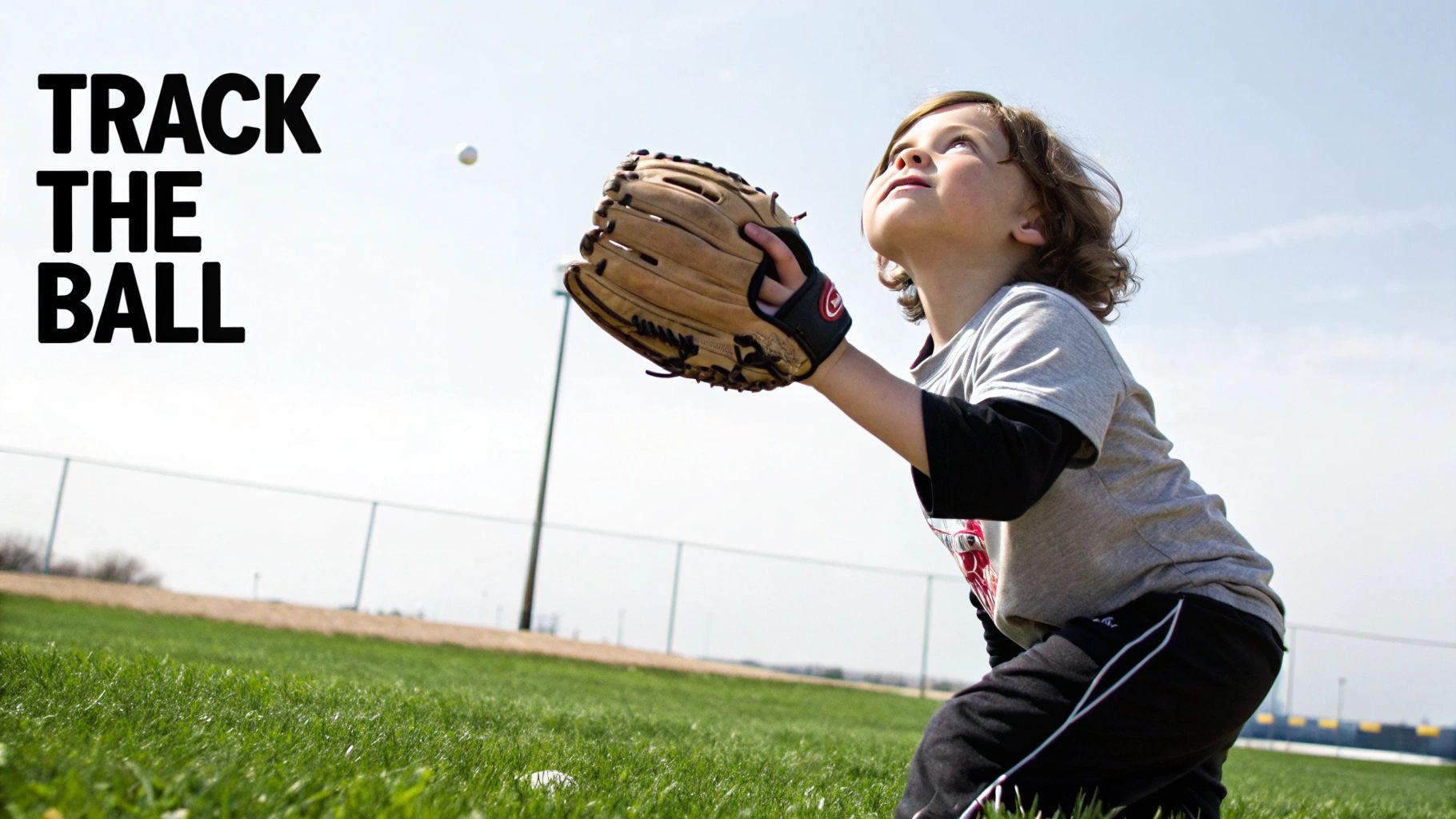 A young child wearing a baseball glove looks up, tracking a baseball in the sky on a sunny day.