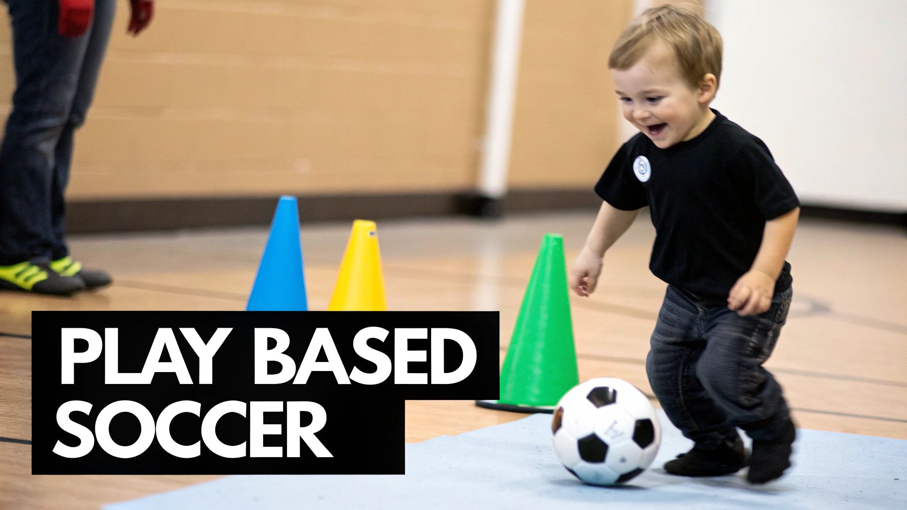 A joyful toddler kicks a soccer ball indoors, surrounded by colorful training cones and an instructor.