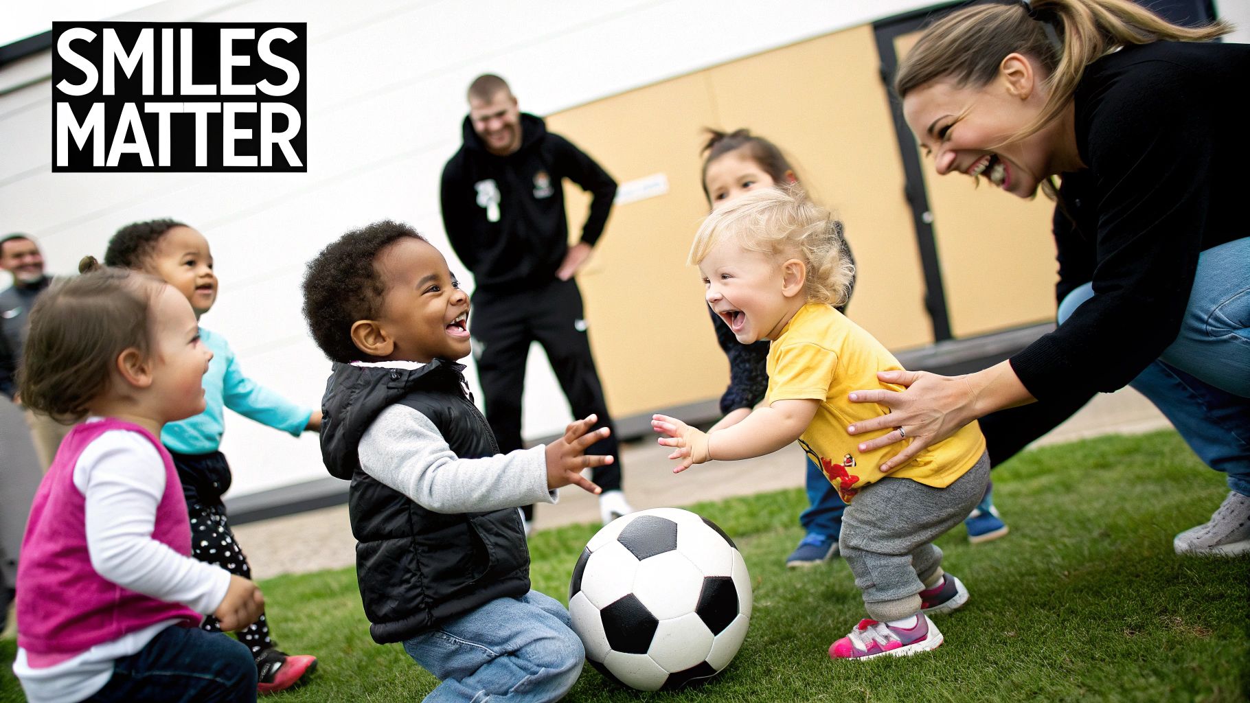 Happy toddlers and adults laughing while playing with a soccer ball on grass.