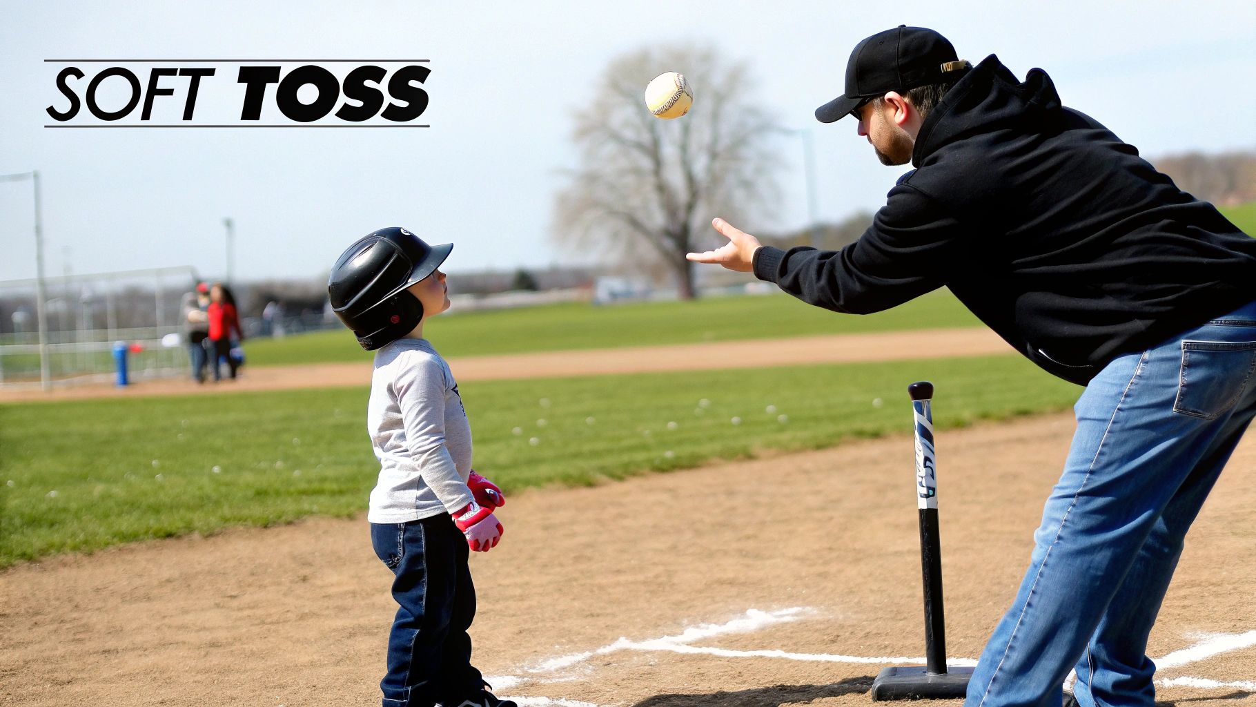 A child in a batting helmet watches a parent or coach soft toss a baseball on a field.