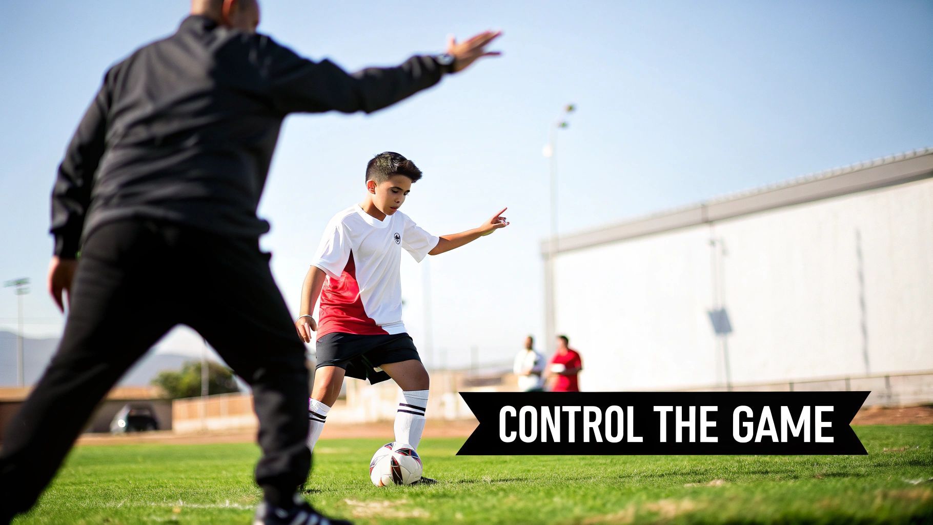 Young soccer player practices ball control on a sunny field with a blurred coach providing guidance.