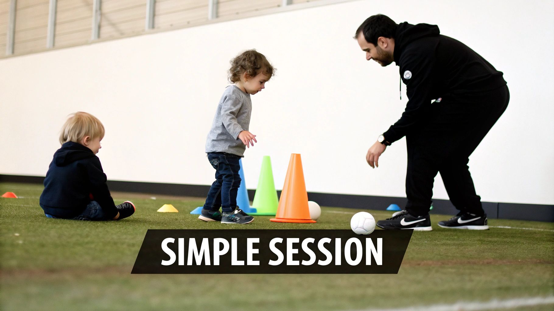 A man coaches two young toddlers on an indoor green turf field with colorful cones and soccer balls.