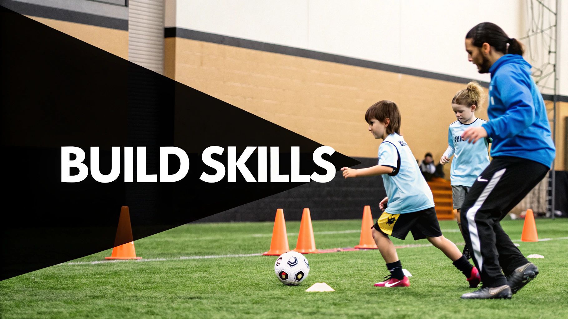 A soccer coach teaches young children to dribble a ball on an indoor turf field with cones.