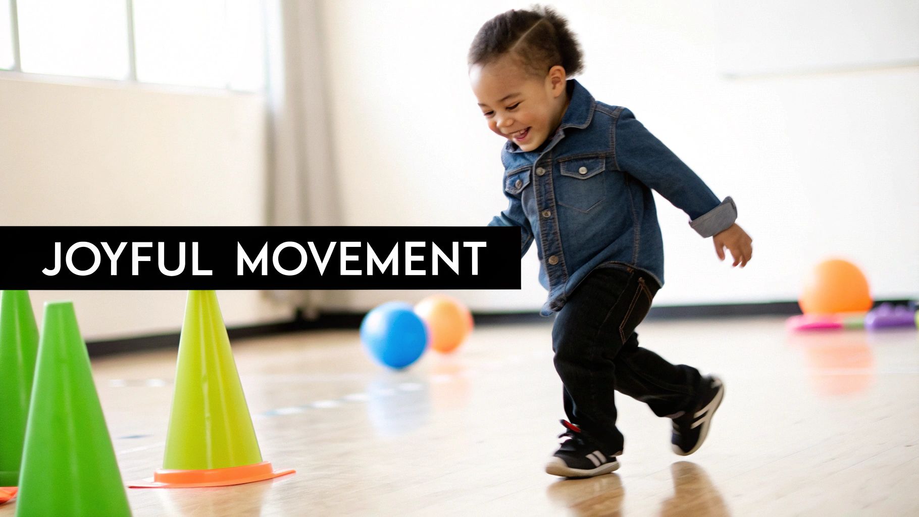A joyful toddler with dark curly hair runs past green cones and colorful balls on a wooden floor.