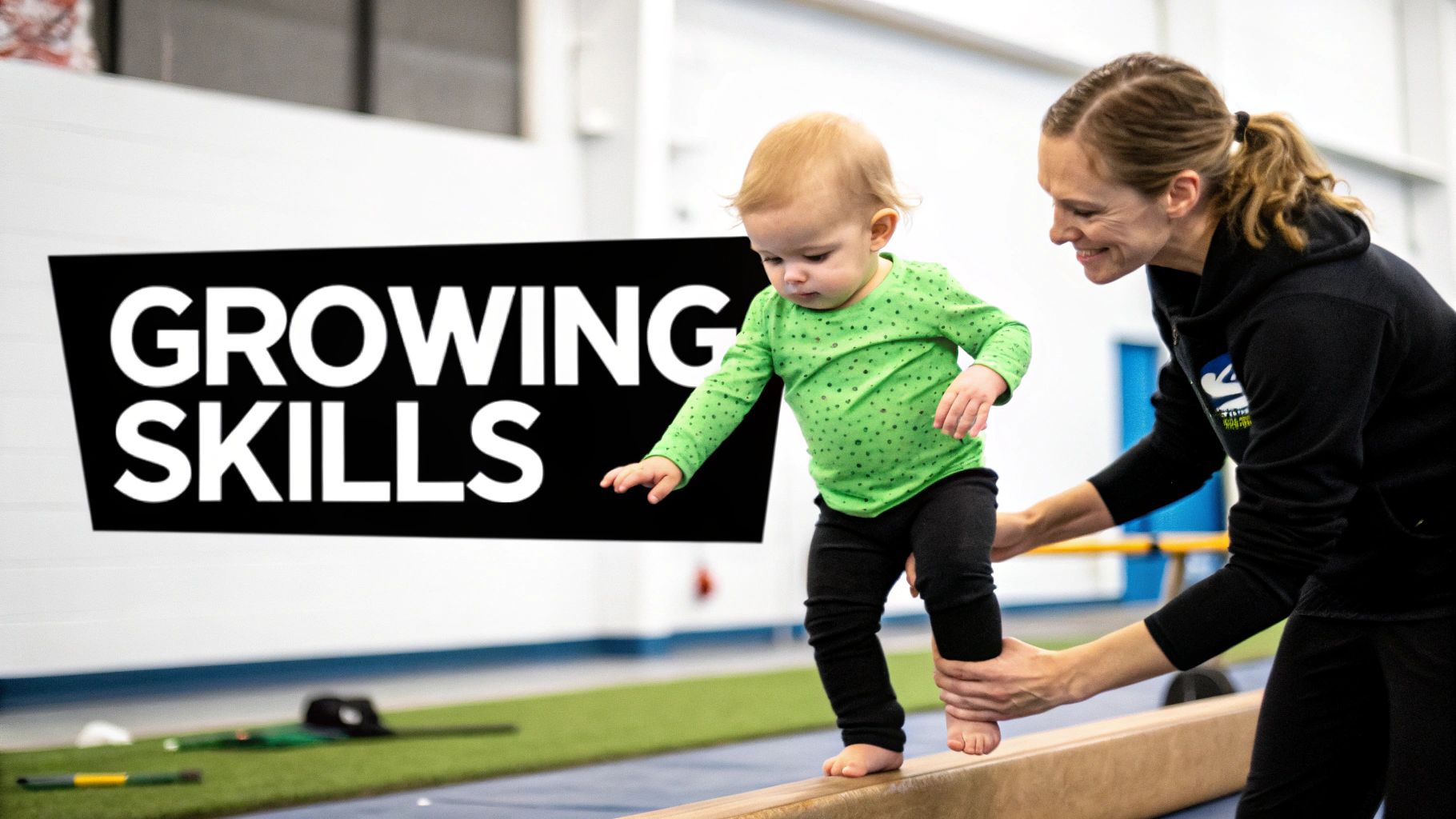 A smiling woman helps a toddler in a green shirt walk on a balance beam in a gym.